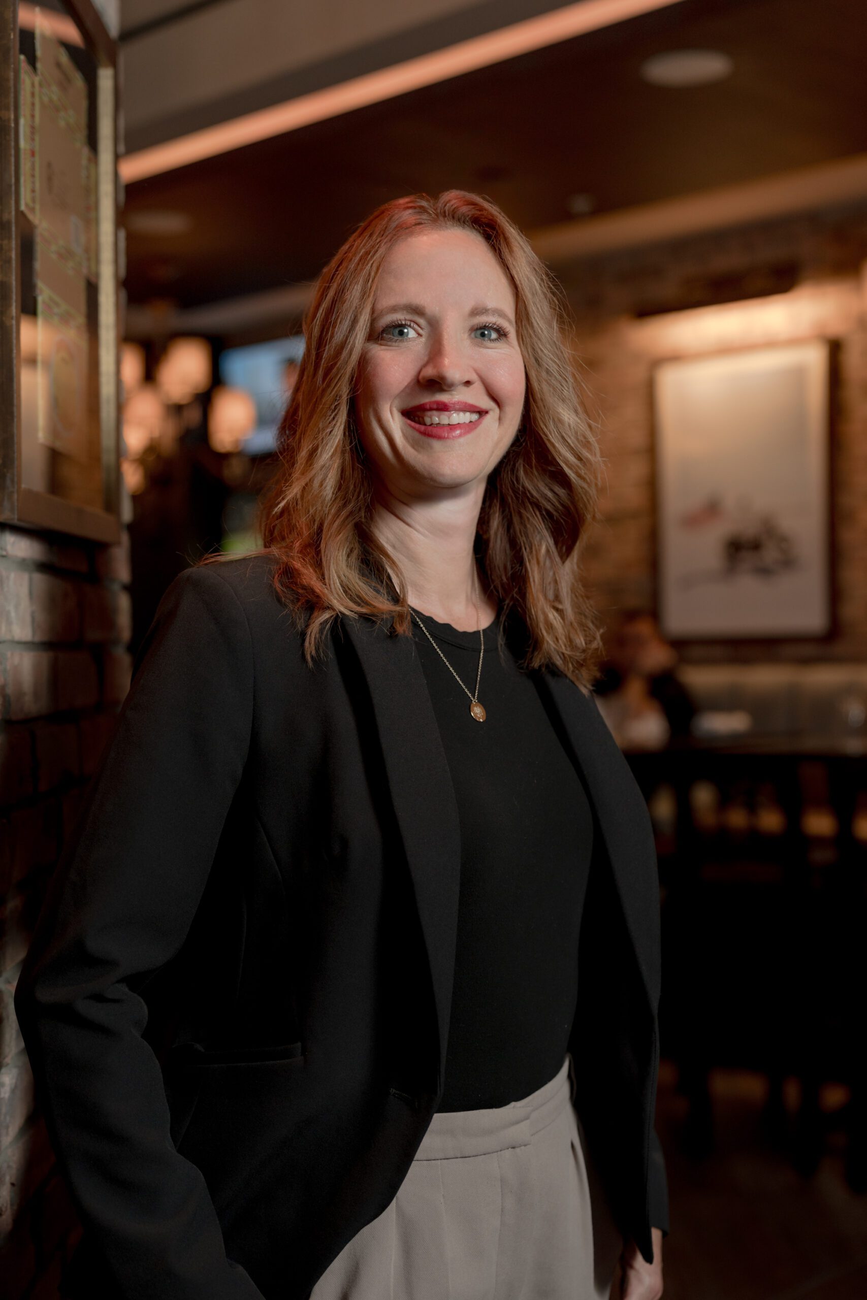 A woman with light brown hair wearing a black blazer and beige pants smiles while standing indoors in a warmly lit, modern restaurant.