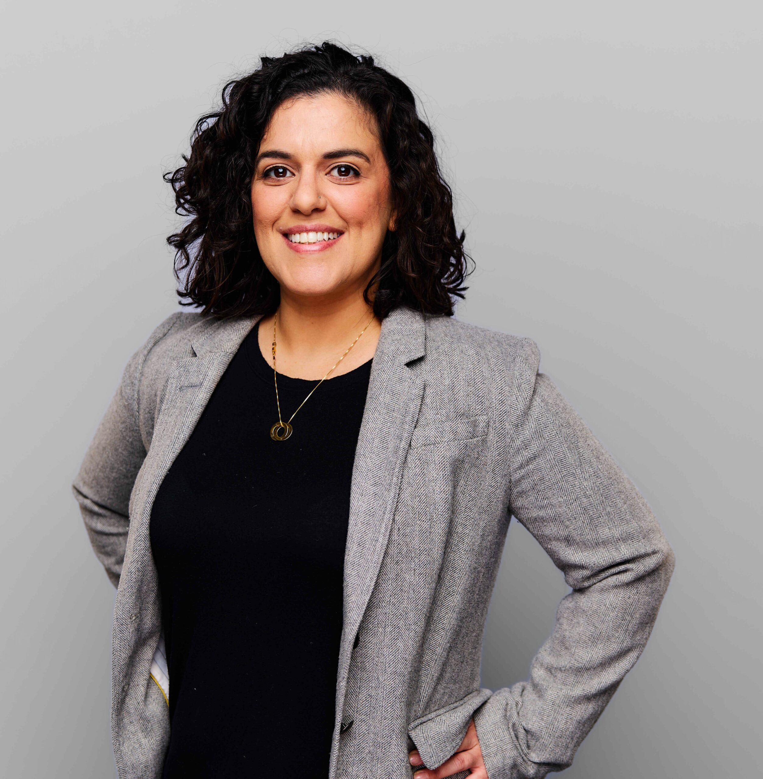 A woman with curly dark hair smiles at the camera, wearing a gray blazer over a black top and a gold necklace. She stands against a plain light gray background with her hands on her hips.
