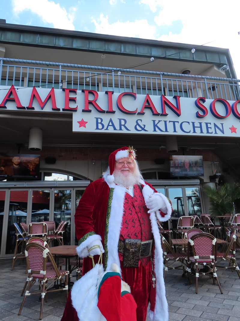 A person dressed as Santa Claus stands smiling in front of an outdoor bar and kitchen with empty tables and chairs, ready to serve festive food and beverage under a sign that reads 