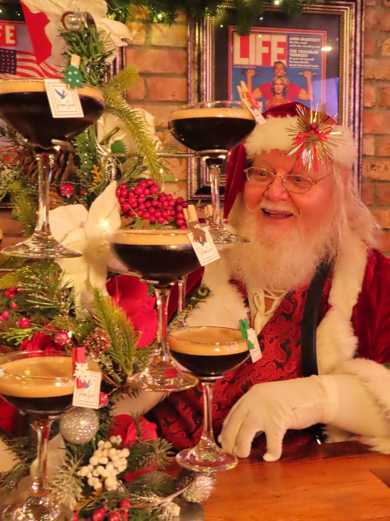Santa Claus smiles beside a festive arrangement of espresso martinis stacked in coupe glasses, adorned with holiday greenery and mini ornaments—perfect for celebrating both the season and upcoming Halloween events, with a framed LIFE magazine cover in the background.
