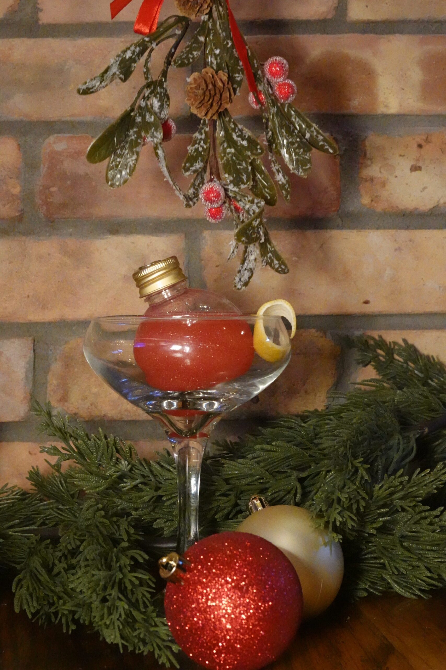 A festive cocktail in a coupe glass with a red ornament-shaped bottle and a lemon twist, surrounded by pine branches, red and gold ornaments—perfect for holiday or Halloween events—under mistletoe with a brick wall backdrop.
