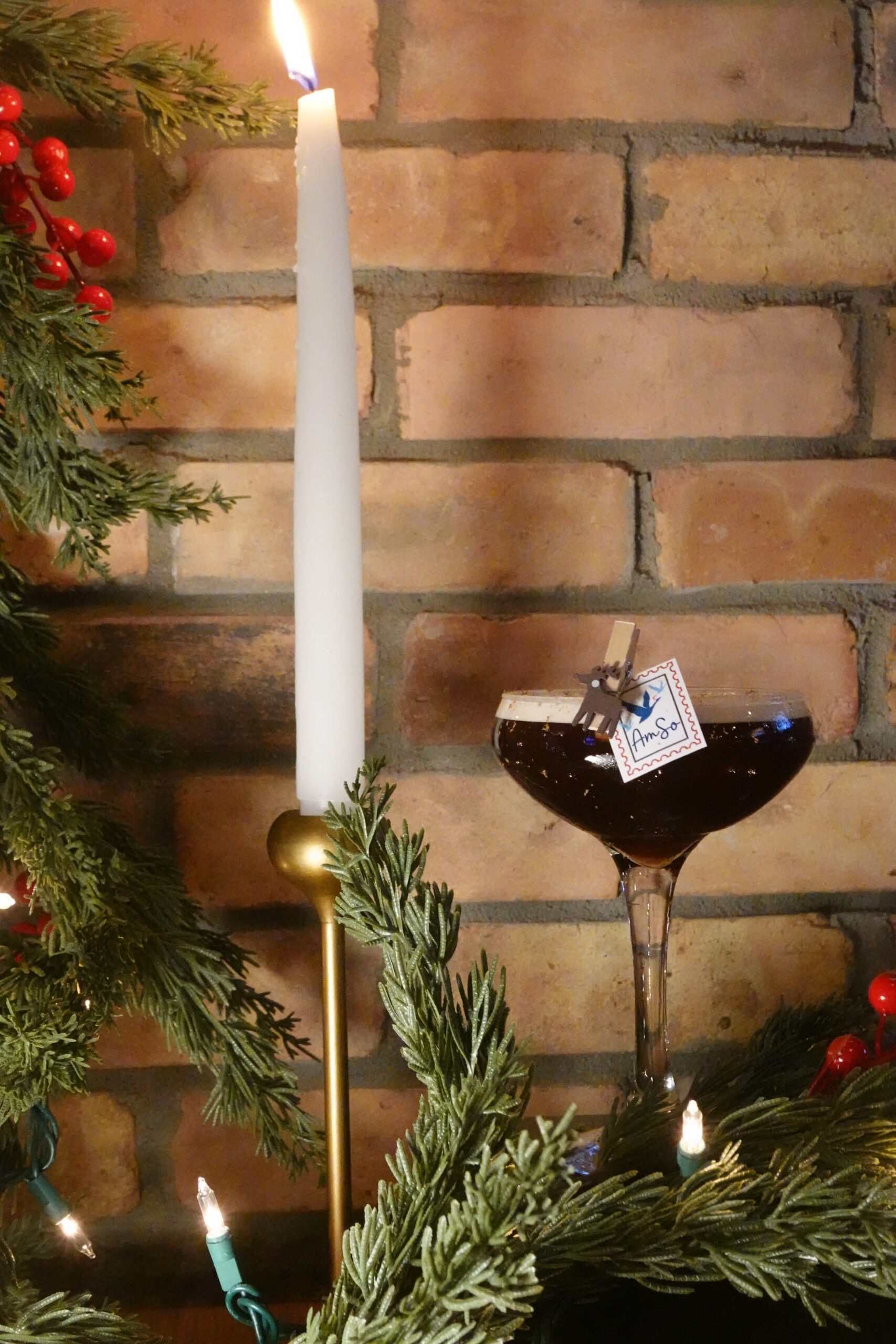 A festive scene with a tall white candle in a gold holder, a cocktail glass filled with a dark drink, and evergreen branches with red berries and string lights, perfect for cozy gatherings or halloween events against a brick wall.