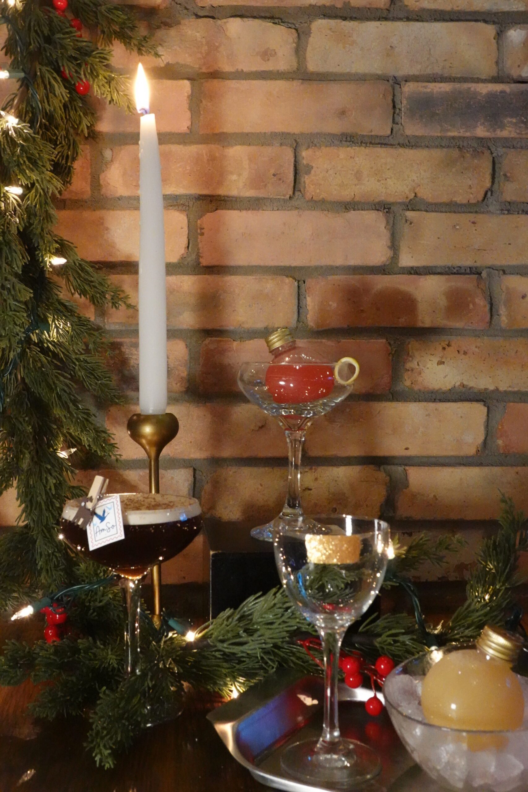Three elegant cocktails in glassware are displayed on a wooden table with evergreen branches and red berries, beside a tall lit candle in a brass holder—an inviting scene perfect for cozy gatherings or Halloween events, all set against a brick wall background.