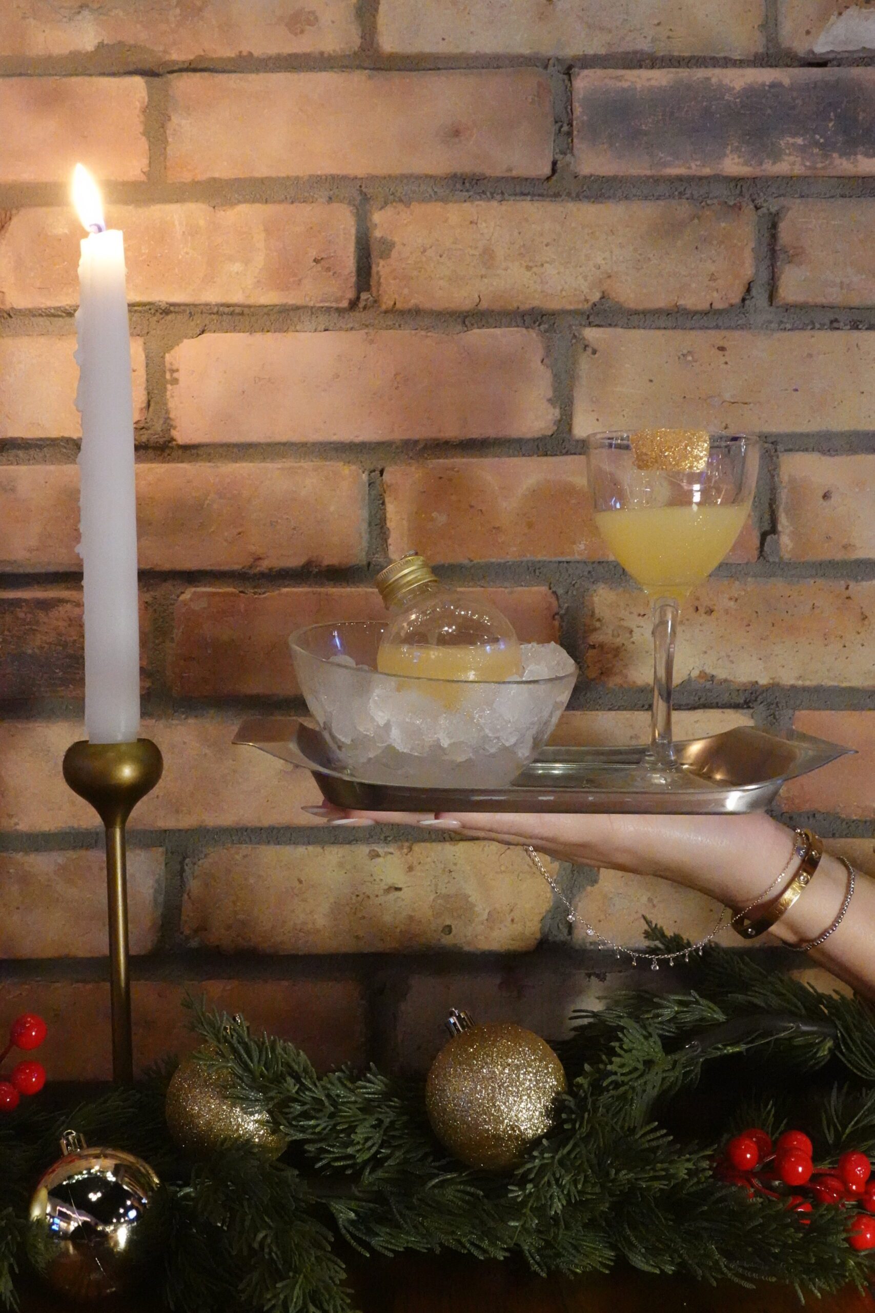 A hand holds a tray with a glass of yellow drink and a bowl of ice cradling a light bulb-shaped bottle, set before a brick wall. A lit candle and festive holiday greenery evoke the cozy magic of Halloween events with gold ornaments.