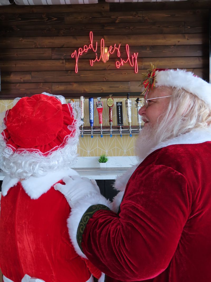 Mr. and Mrs. Claus, dressed in red and white holiday outfits, stand together facing a wall of beer taps beneath a neon sign that reads 