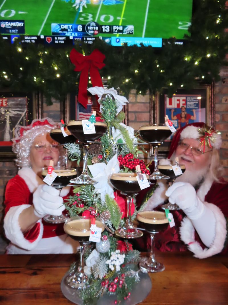 Two people dressed as Mrs. Claus and Santa Claus toast with espresso martinis on a festive holiday glass tower. Sports play on a TV in the background, with Christmas decorations all around—perfect for those who love both holiday cheer and Halloween events.