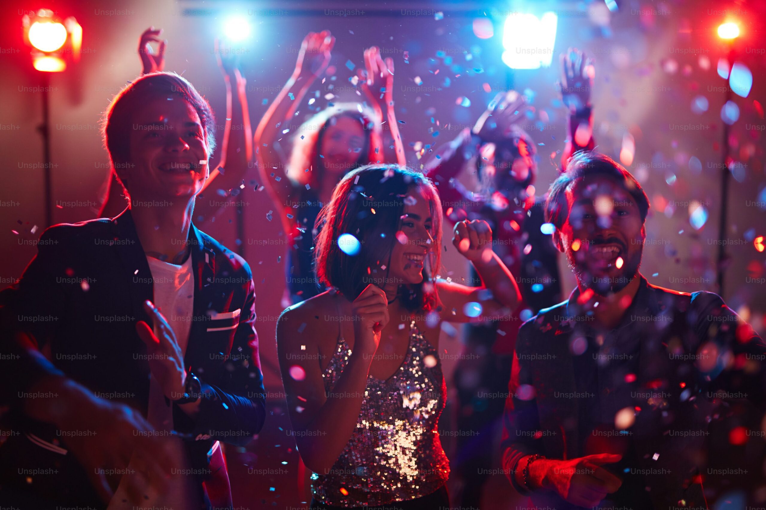 A group of young people dance and celebrate at a New Year’s Eve party, surrounded by colorful lights and falling confetti. They are smiling and appear to be enjoying the lively atmosphere.