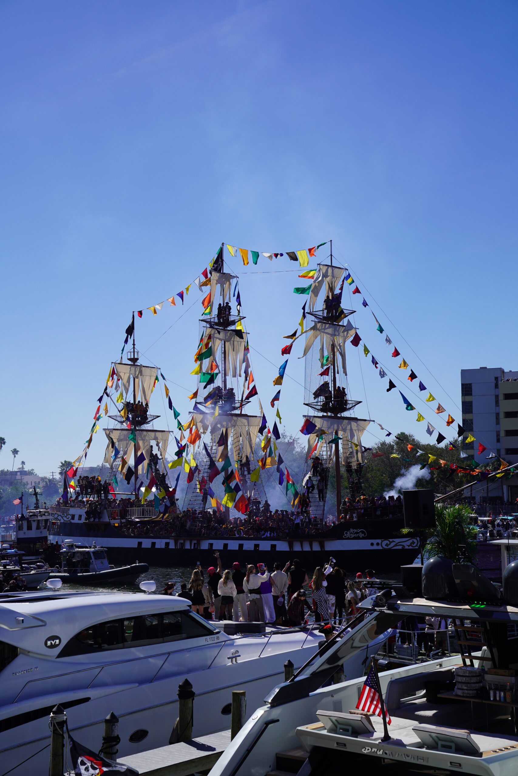 A large, decorated pirate ship with colorful flags sails in a harbor during a festival. People crowd the ship and nearby boats, enjoying festive food and beverage under a bright blue sky as smoke rises from the ship in celebration.