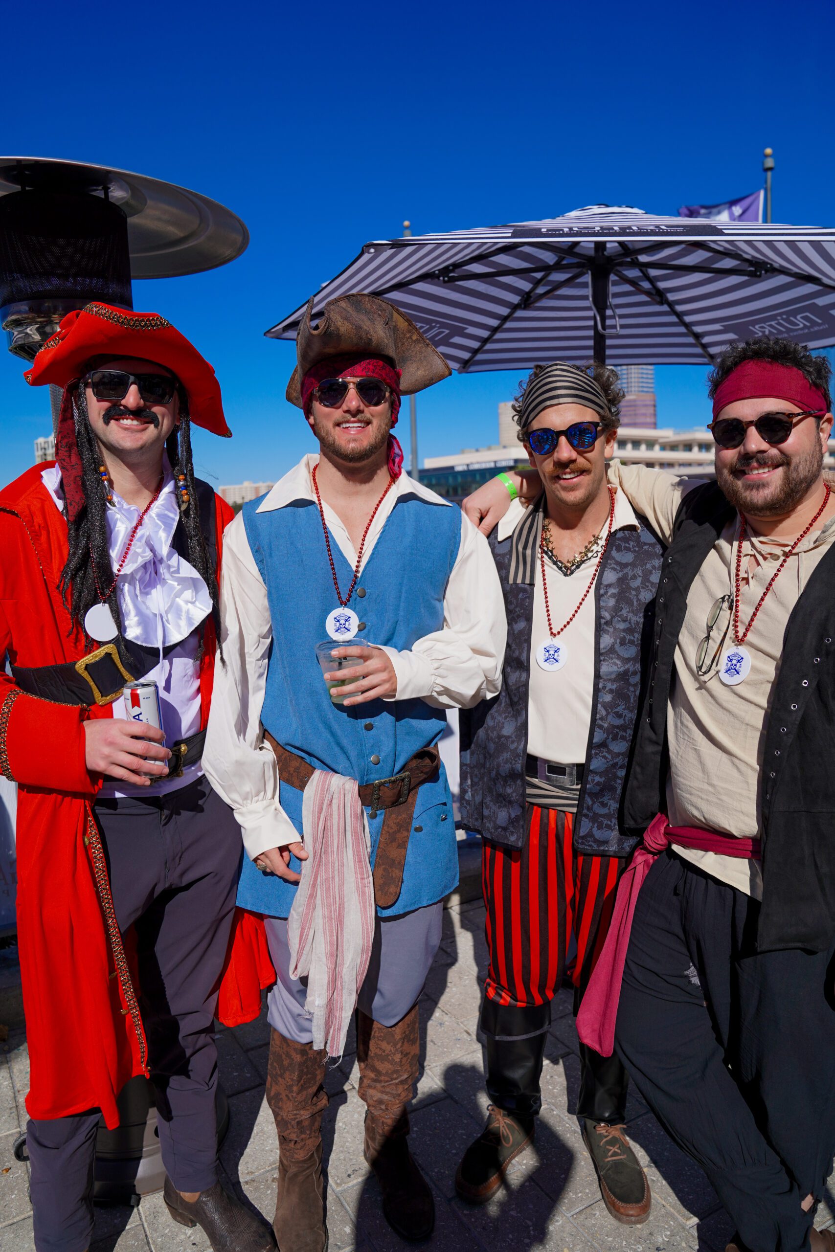 Four men dressed in colorful pirate costumes stand together outdoors on a sunny day, smiling at the camera. With festive food and beverage nearby, they wear hats, sunglasses, and accessories beneath a large umbrella and blue sky.