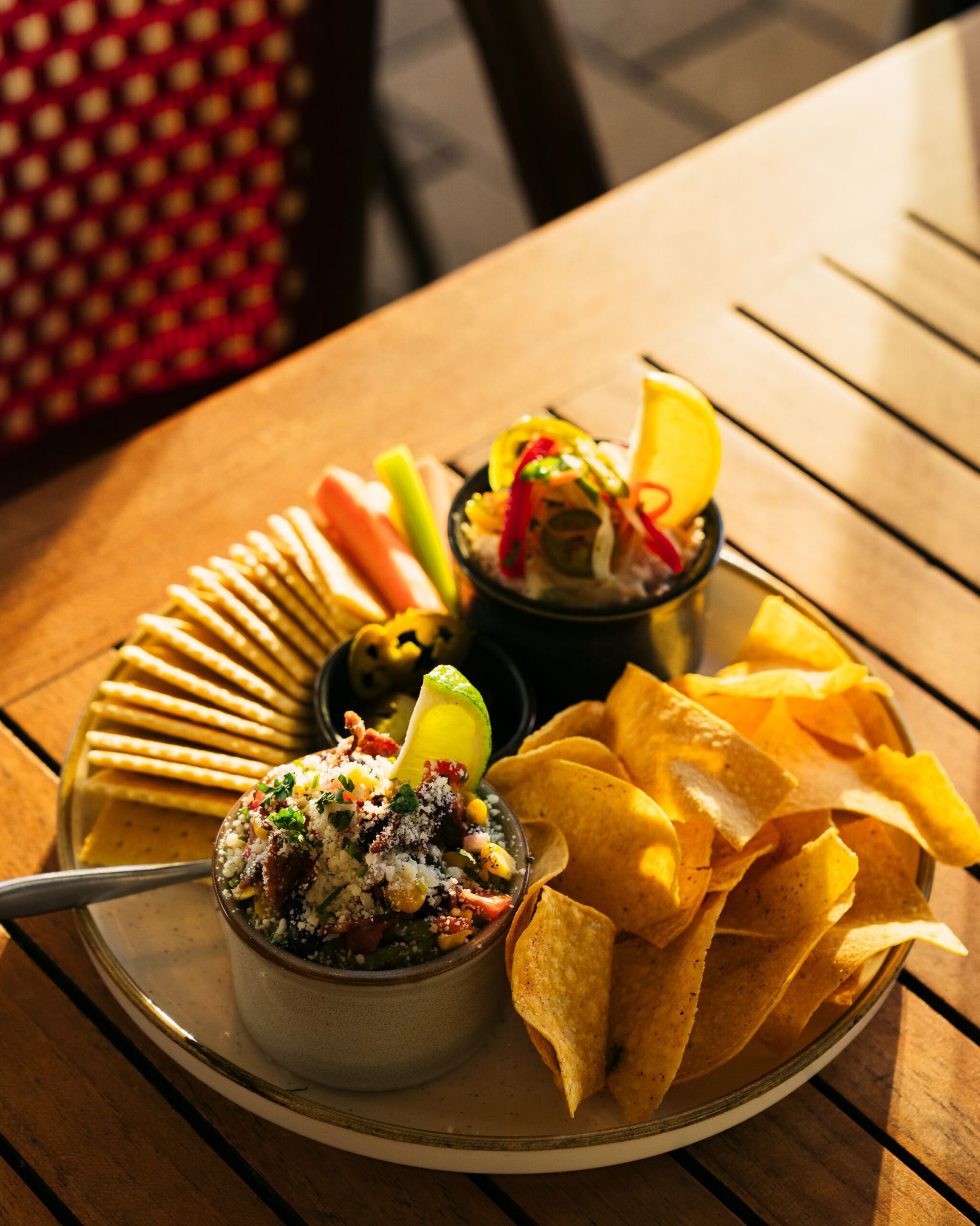 A plate with tortilla chips, round crackers, veggie sticks, two dips garnished with lime, herbs, and sliced vegetables, served on a wooden table in warm sunlight.