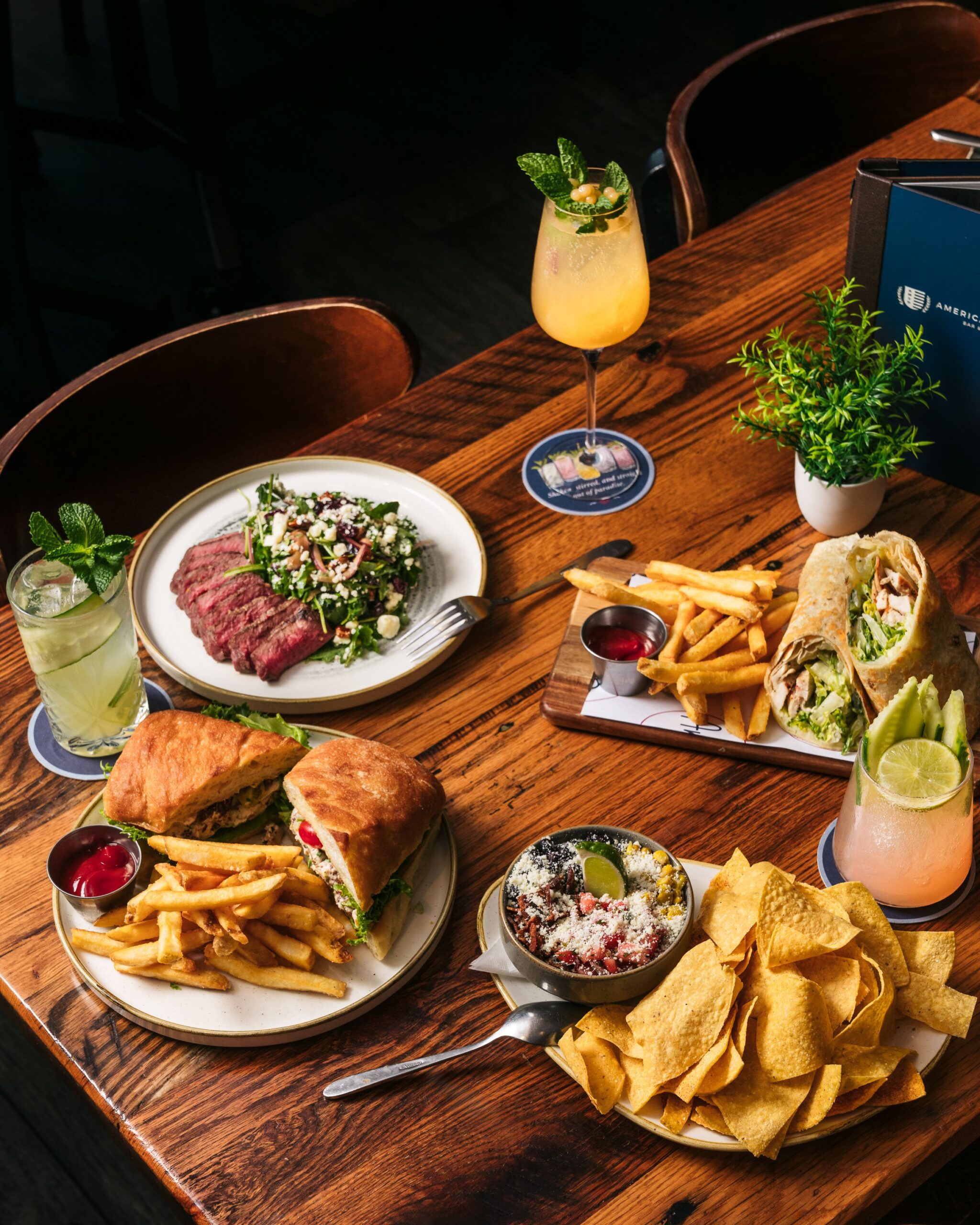 A wooden table at American Social displays plates of food, including a steak salad, chicken wrap with fries, sandwich with fries, tortilla chips with dip, and three colorful drinks garnished with mint and fruit.