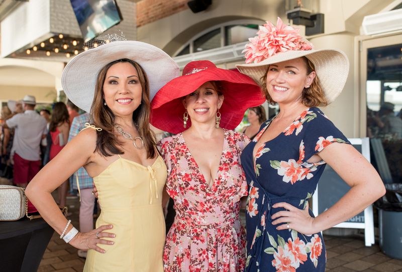 Three women wearing colorful, wide-brimmed hats and floral dresses smile and pose together at the F1 Miami Grand Prix, with other people and festive decorations visible in the background.