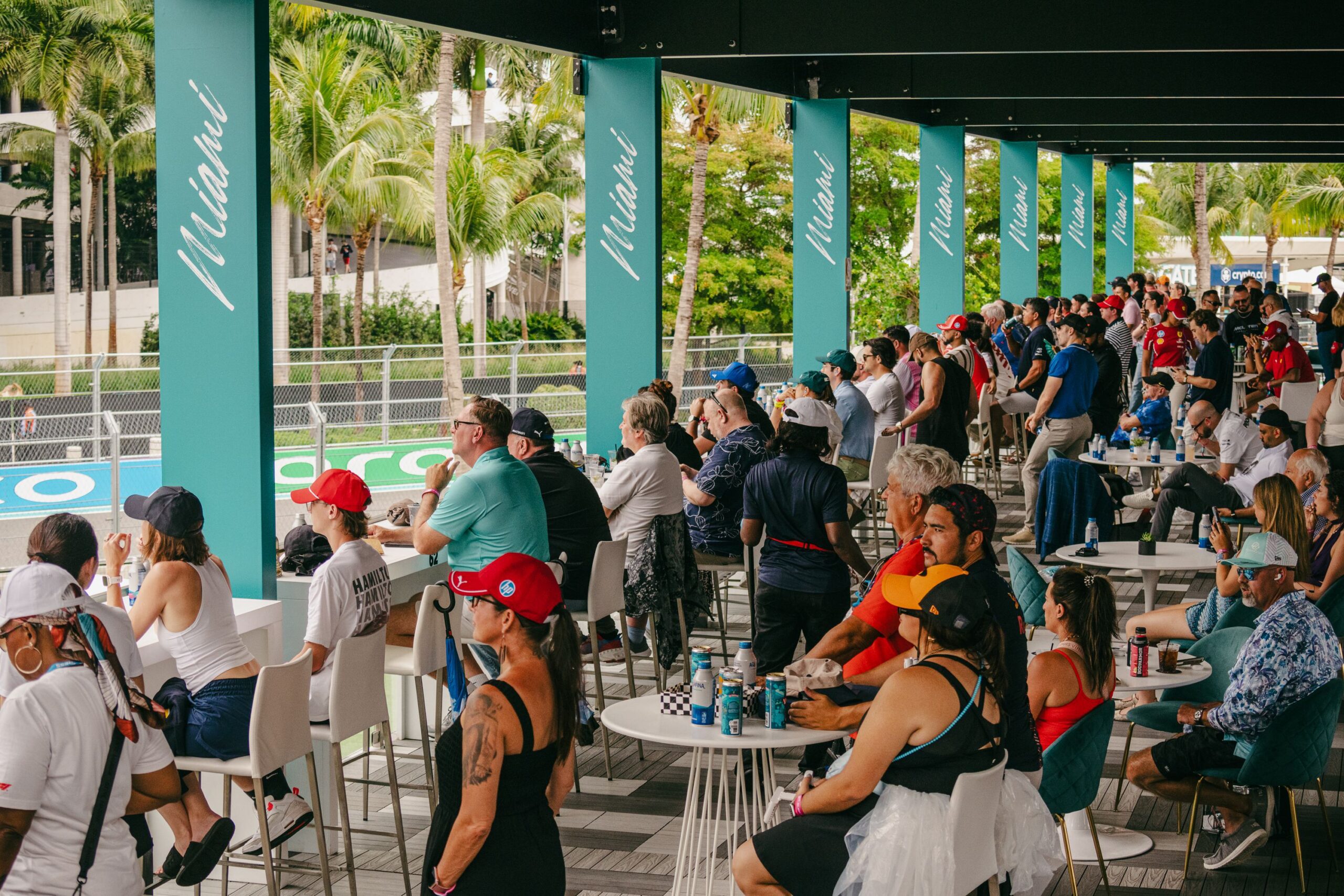 A large crowd of people, many wearing hats, sit and stand on a covered terrace overlooking the racetrack at the F1 Miami Grand Prix, with palm trees and 