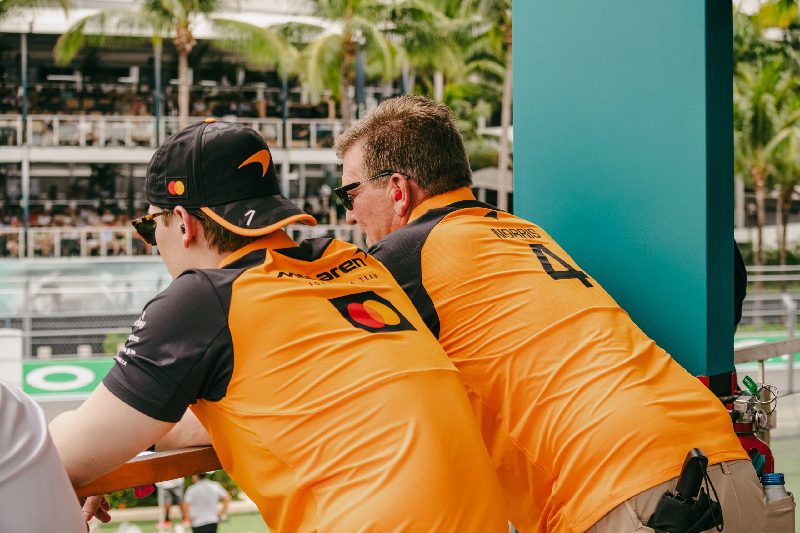 Two people in orange and black McLaren team shirts lean on a railing, watching the F1 Miami Grand Prix. Palm trees and spectators fill the stands in the background.