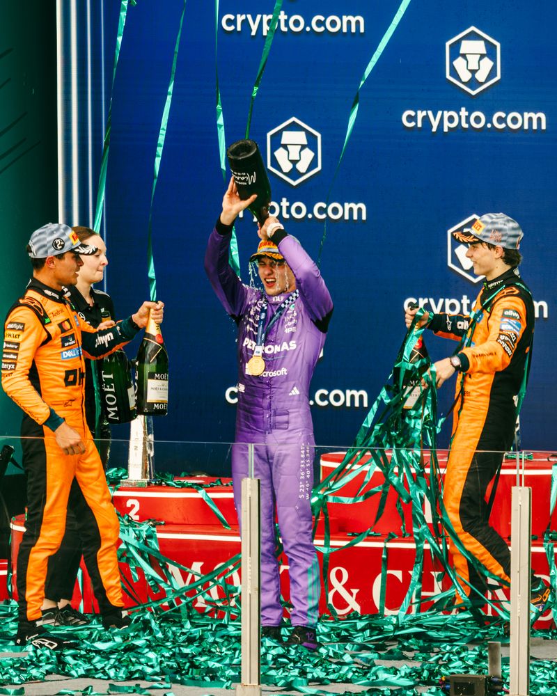 Three race car drivers celebrate on the podium at the F1 Miami Grand Prix with champagne and green streamers; the center driver in purple holds a bottle overhead, flanked by two in orange and black, as a blue 