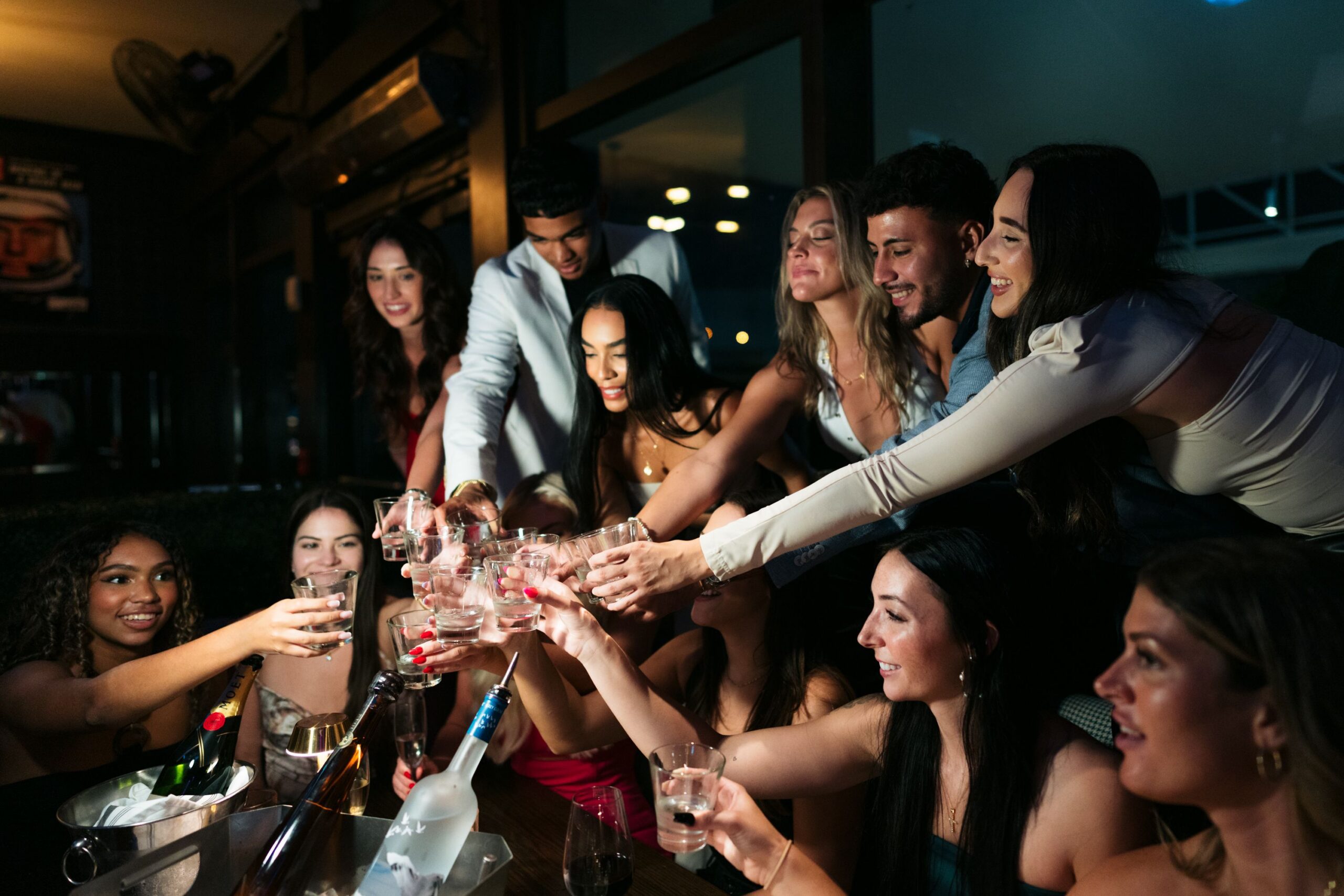 A group of young adults dressed up and smiling clink their glasses together while celebrating at a dimly lit, boozy restaurant or bar, with bottles and drinks visible on the table in front of them.