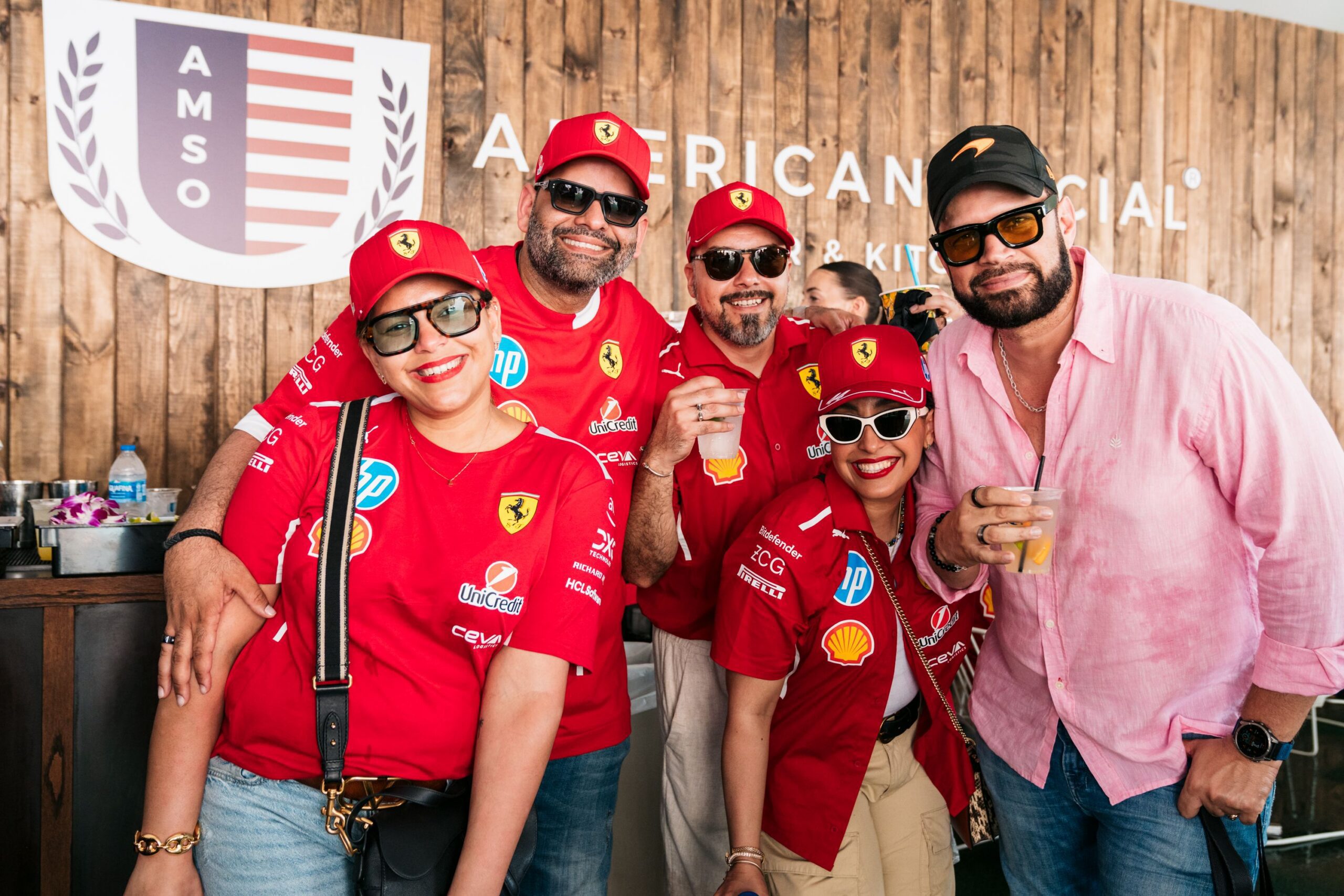 Five smiling people pose together, four in red Ferrari shirts and caps, one in a light pink shirt and cap. In front of a wooden wall with a logo and text, they enjoy a Late Night gathering as one person holds a drink.
