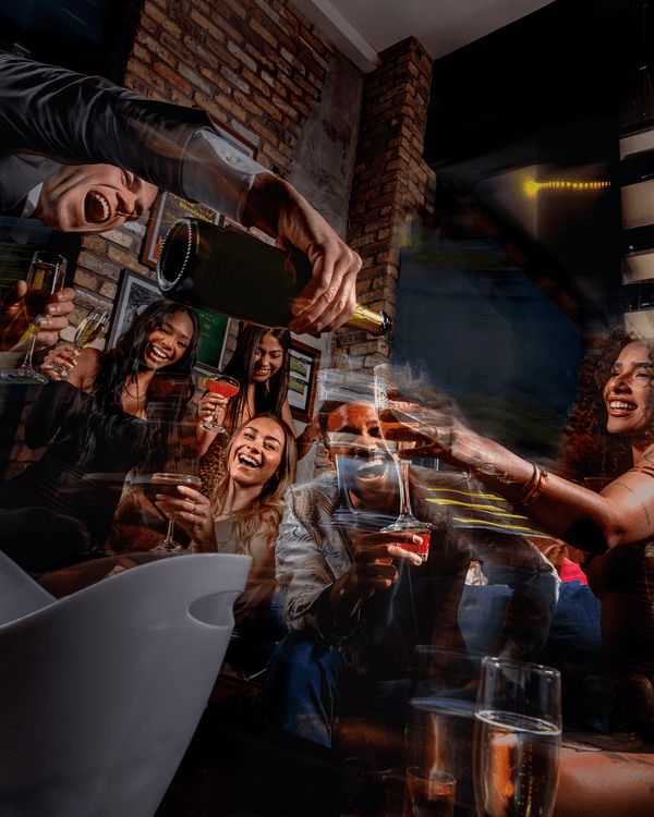 A group of friends are laughing and raising glasses in a lively indoor setting as one person pours champagne, creating a boozy, celebratory atmosphere with festive energy and exposed brick walls in the background.
