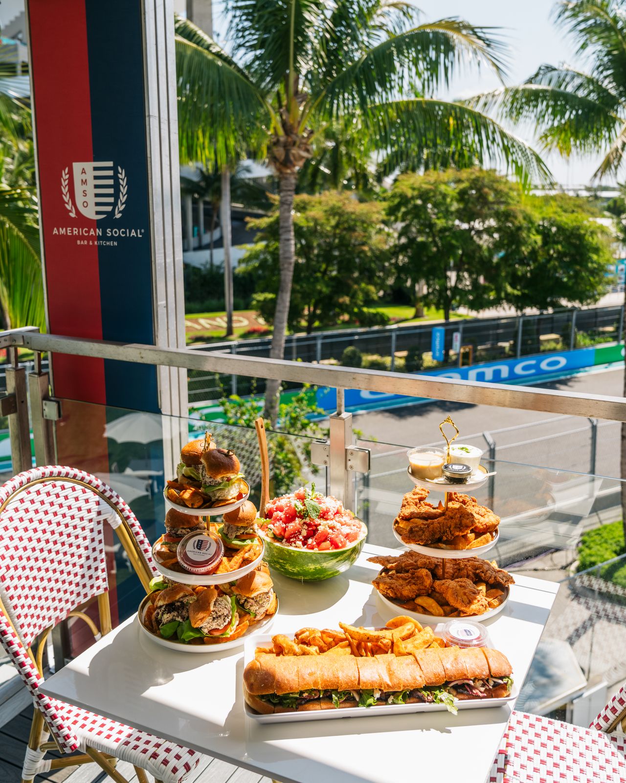 A table on a sunny balcony is filled with burgers, fried chicken, sandwiches, and a bowl of salad. Palm trees and race track barriers hint at the F1 Miami Grand Prix nearby. The scene is vibrant and inviting.