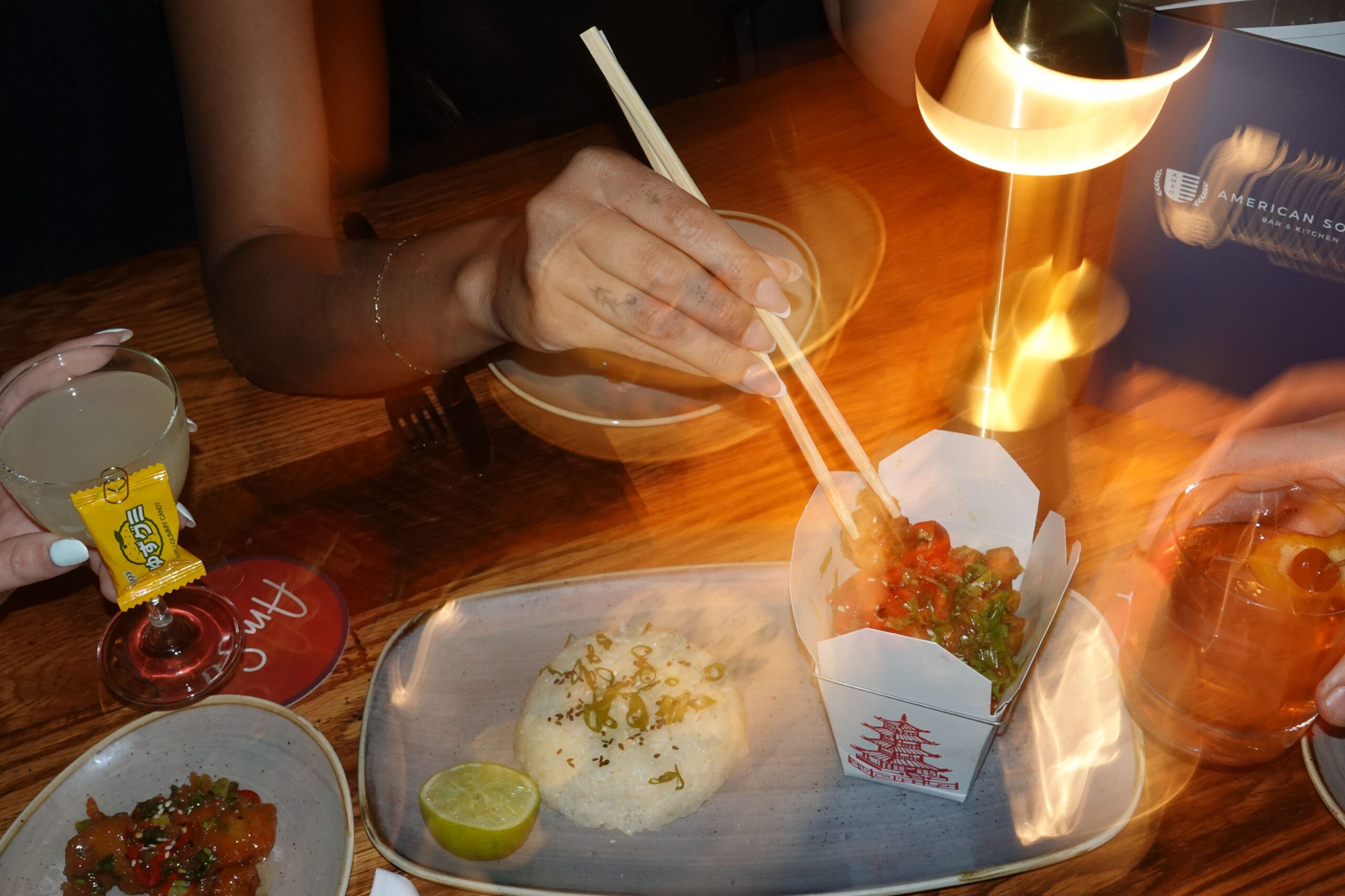 A person eats from a white takeout container with chopsticks at a restaurant table, surrounded by rice, lime, and other dishes. A boozy drink, lamp, and menu are visible, with motion blur capturing the lively hand movement.