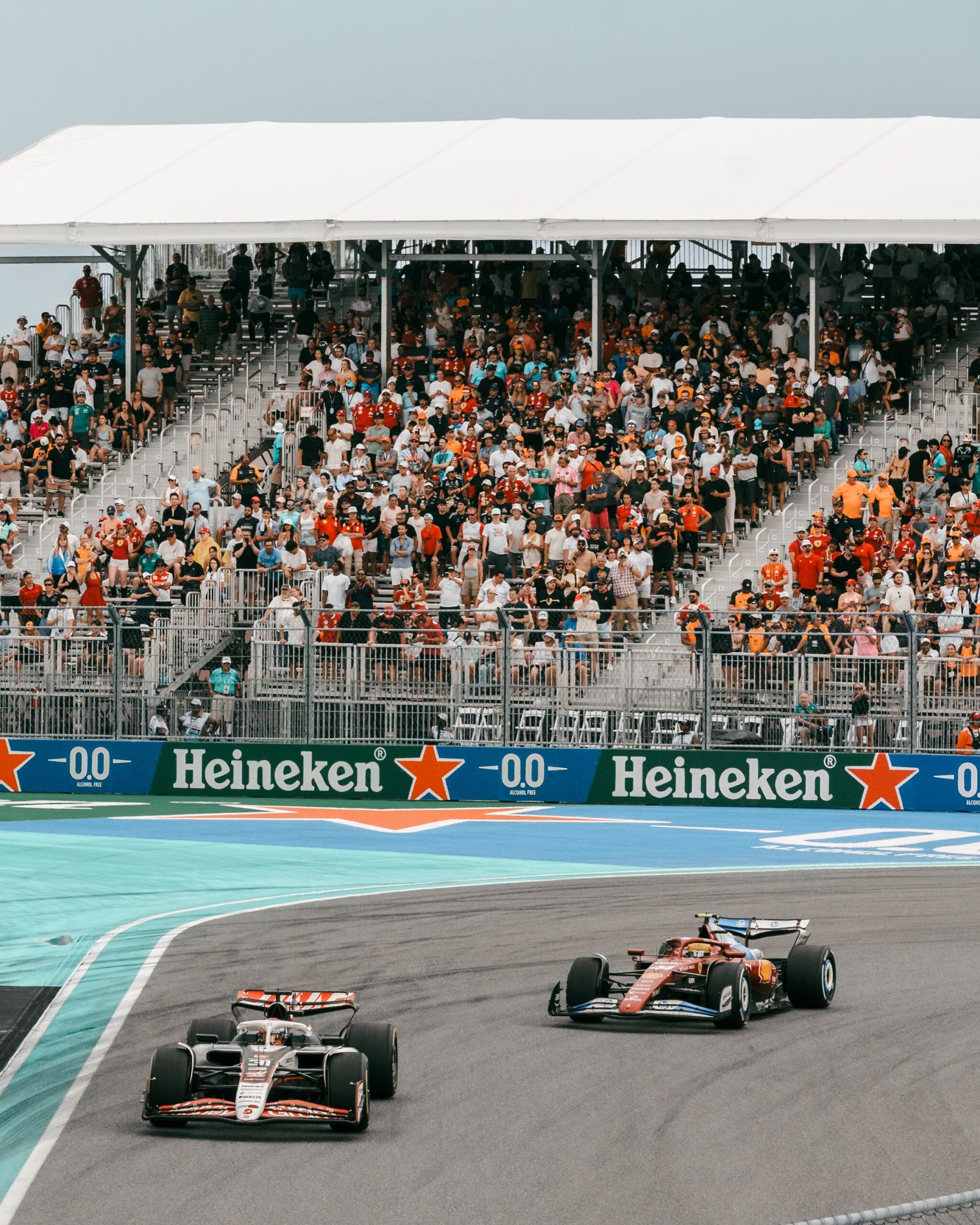 Two Formula 1 cars race on a track near a bend under the glow of late night lights, with a grandstand full of spectators in the background and Heineken advertisements along the barrier.