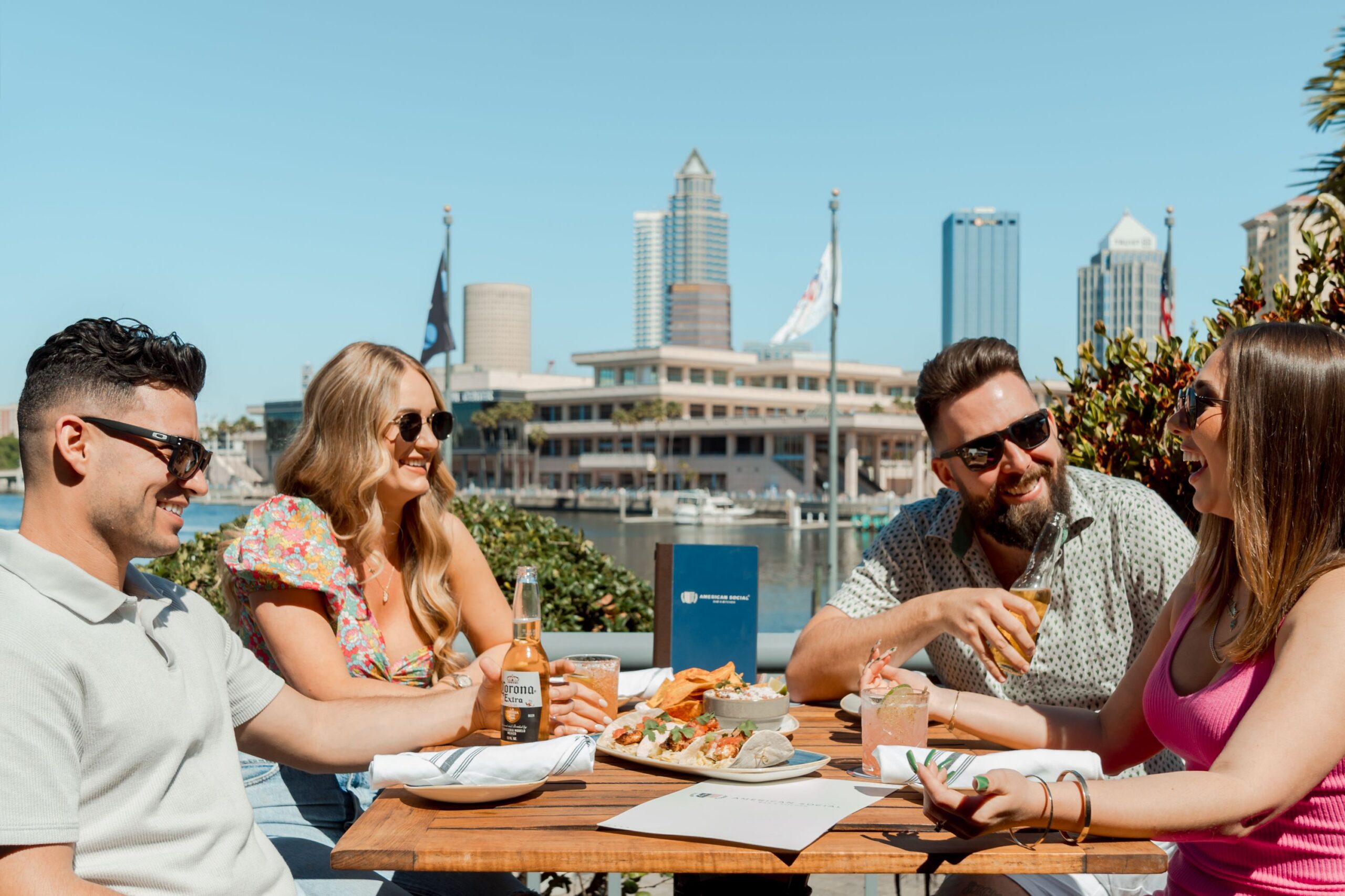 Four friends sit at an outdoor restaurant table, smiling and enjoying drinks and food on a sunny day with views of the city skyline, water, and the excitement of the F1 Miami Grand Prix in the background.