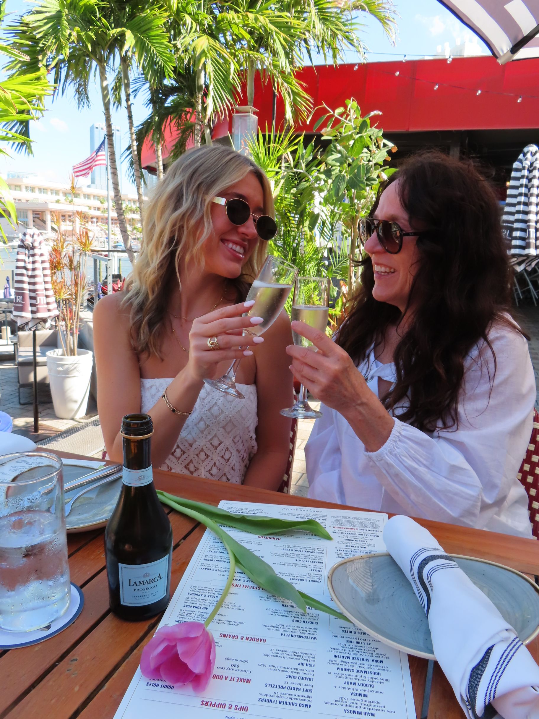 Two women sit at an outdoor restaurant table during the F1 Miami Grand Prix, smiling and clinking champagne glasses. One wears white and sunglasses; the other has long dark hair. A pink tulip, menus, palm trees, and sunlight set the scene.
