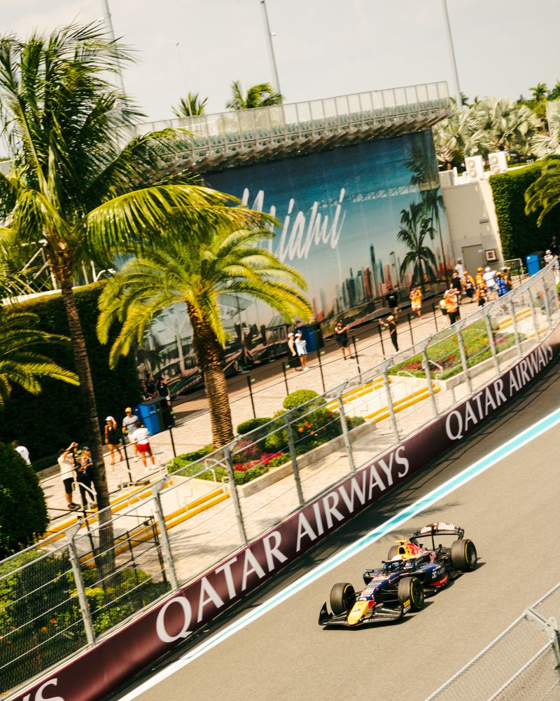 A Formula 1 car races on a track lined with palm trees and Qatar Airways banners at the F1 Miami Grand Prix. Spectators stand behind barriers, with a large 