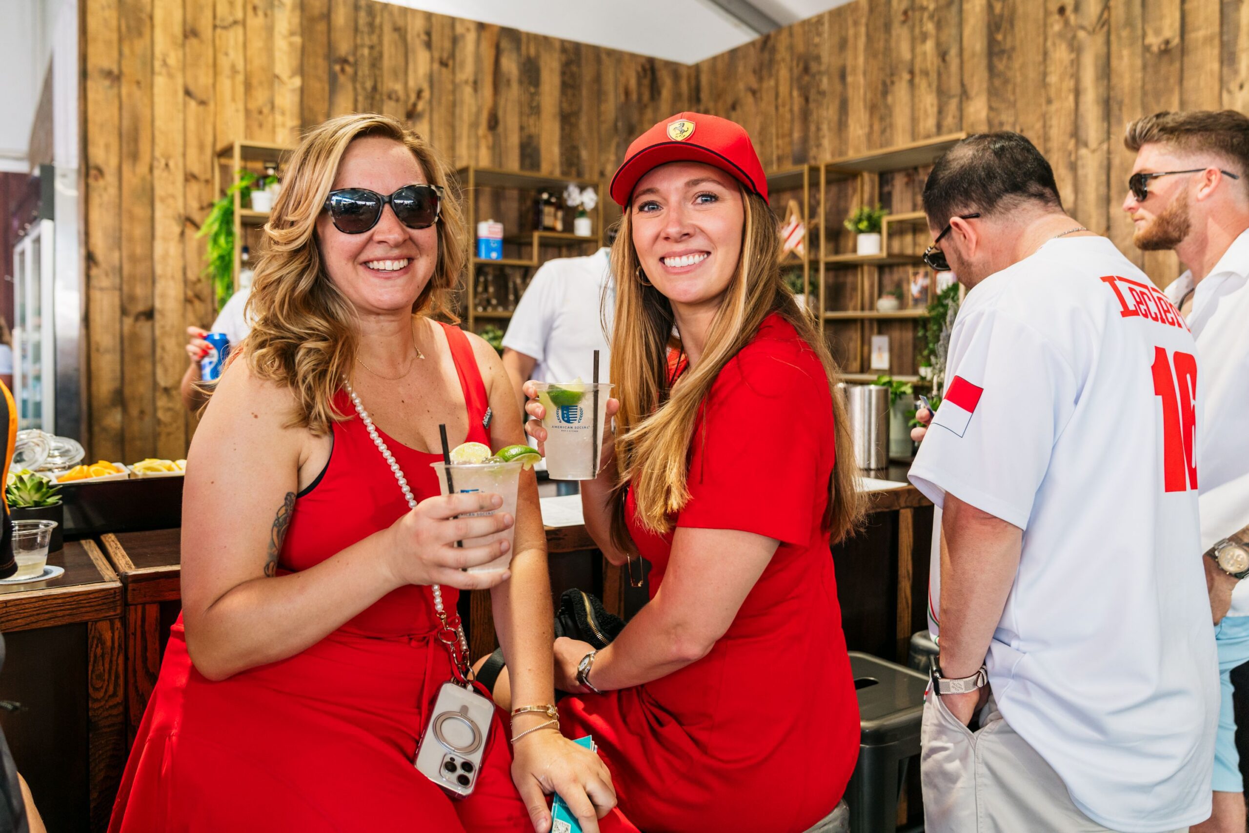 Two women in red dresses smile and hold drinks at an outdoor bar during a Late Night gathering. One wears sunglasses, the other a red cap. Nearby, people—including a man in a white jersey—mingle against wood paneling and drink supplies.