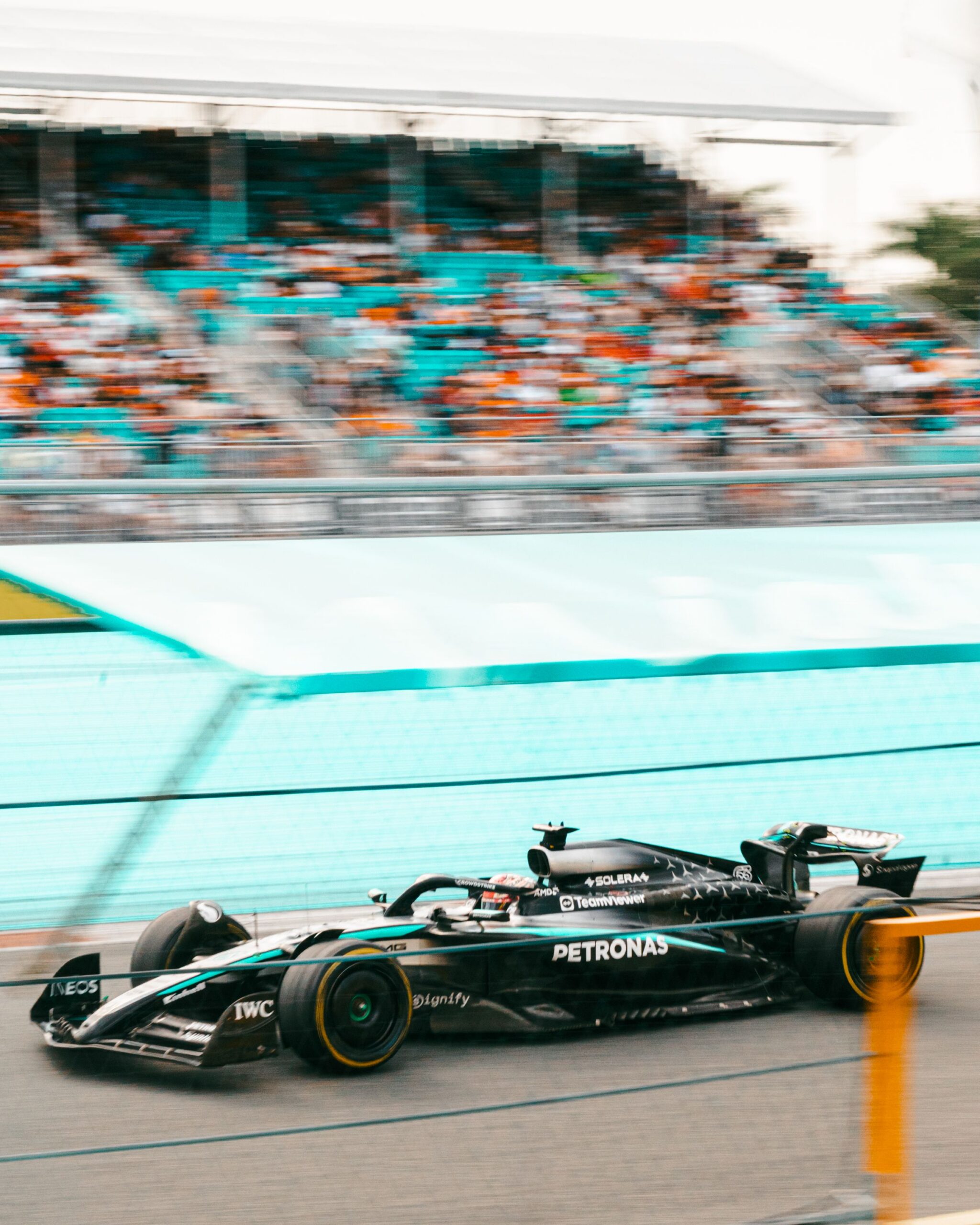 A black Formula 1 Mercedes race car speeds past blurred grandstands full of spectators during a late night race, with motion blur emphasizing its fast movement.