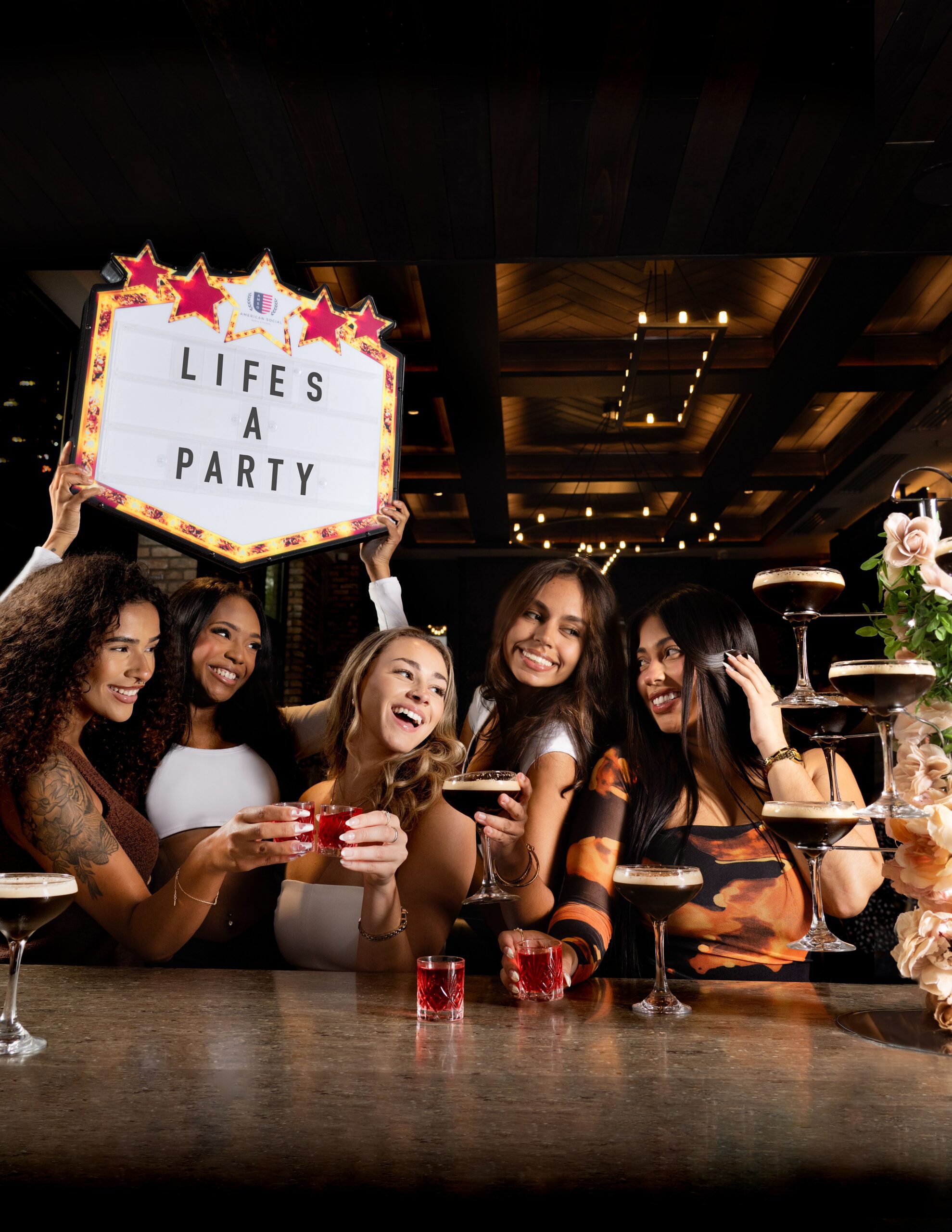 Five women at a bar celebrate together; one holds up a lighted sign reading 