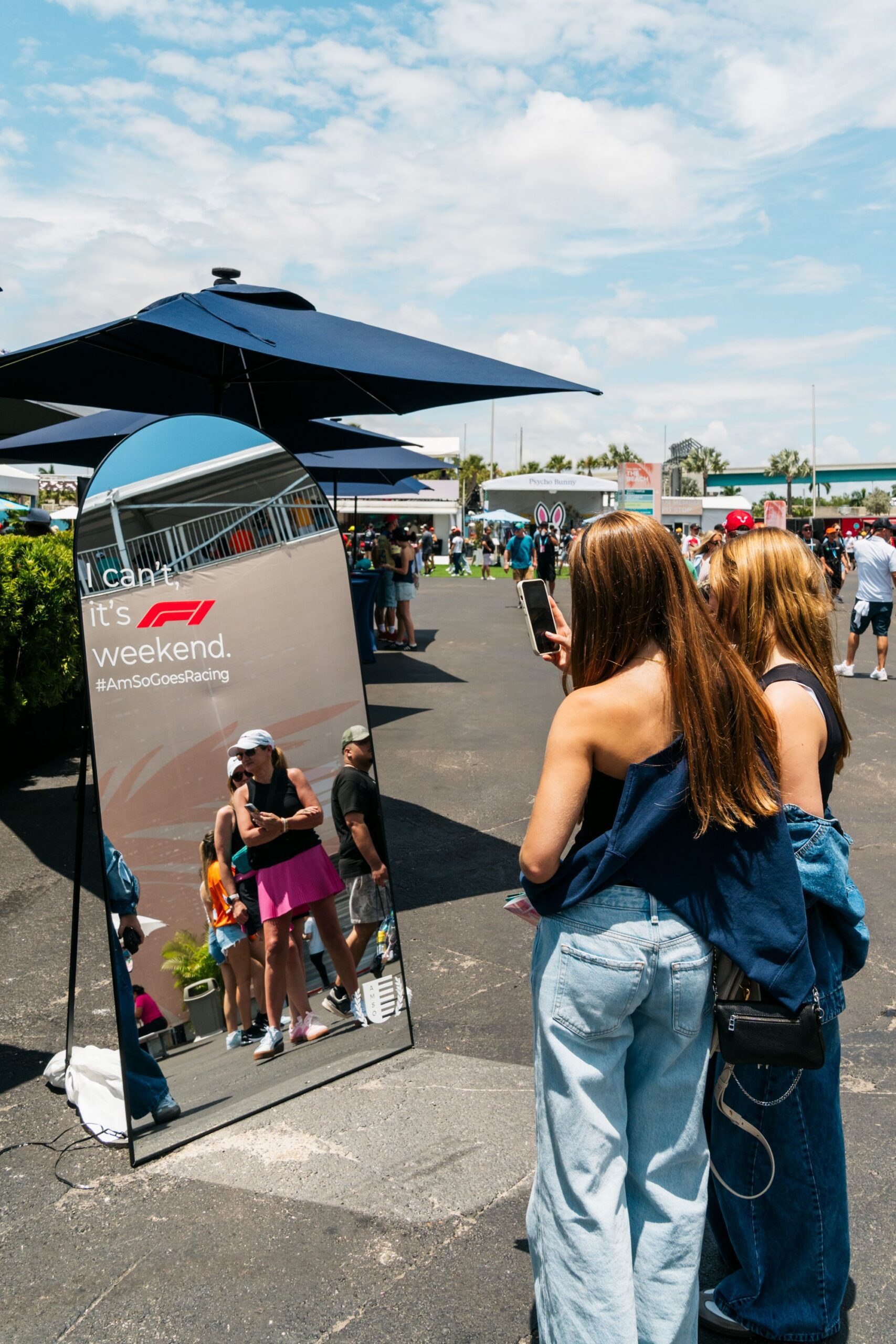 Two women stand outdoors, taking a selfie in a tall mirror with the text “I can. It’s F1 weekend. #AmsSoGoesRacing,” celebrating the excitement of the F1 Miami Grand Prix. People and event tents are visible under a sunny sky.