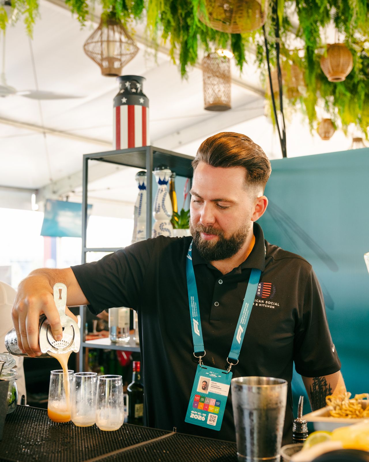 A bearded bartender in a black polo shirt pours cocktails into glasses at a bar, surrounded by greenery and festive decorations, capturing the vibrant energy of the F1 Miami Grand Prix in a bright, lively setting.