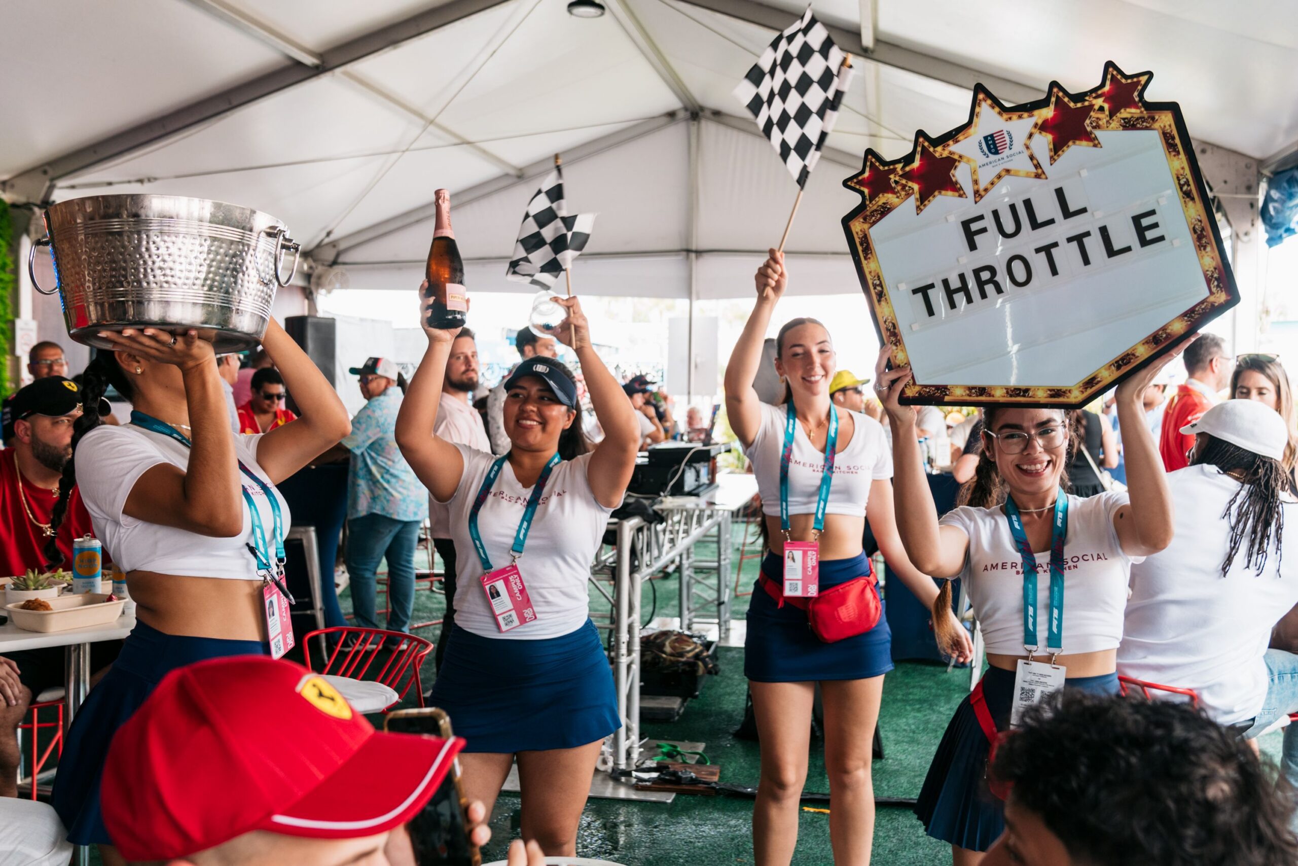 A group of smiling women in matching skirts and white tops celebrate a Late Night event, holding a trophy, champagne bottle, checkered flags, and a 