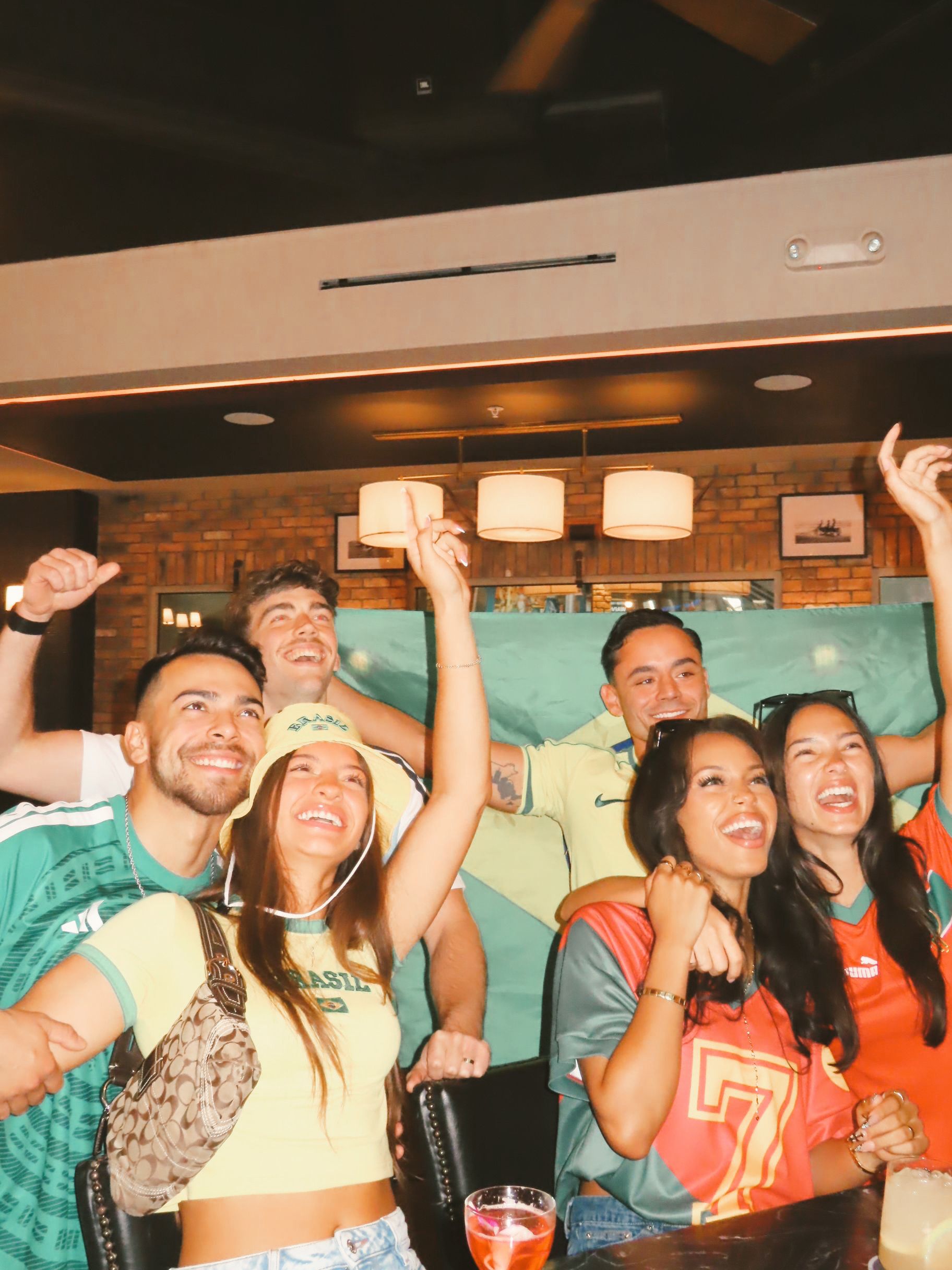 A group of six young adults wearing colorful soccer jerseys smile and cheer together in a bar, with a green flag in the background. They appear excited and joyful, likely celebrating the upcoming World Cup 2026.