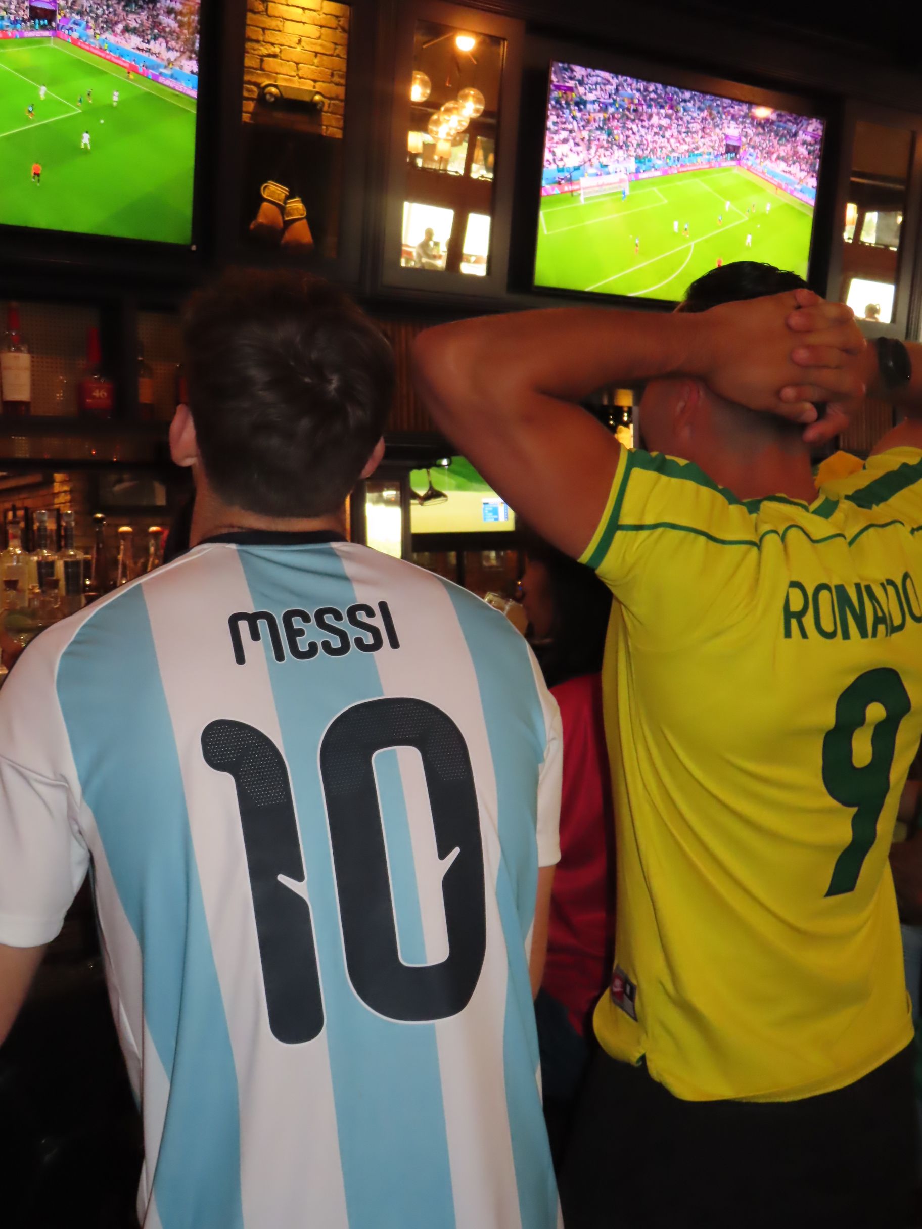 Two people in a bar during Gasparilla watch a soccer match on TV. One wears an Argentina Messi 10 jersey, the other a Brazil Ronaldo 9. The festive atmosphere buzzes with excitement and tension.