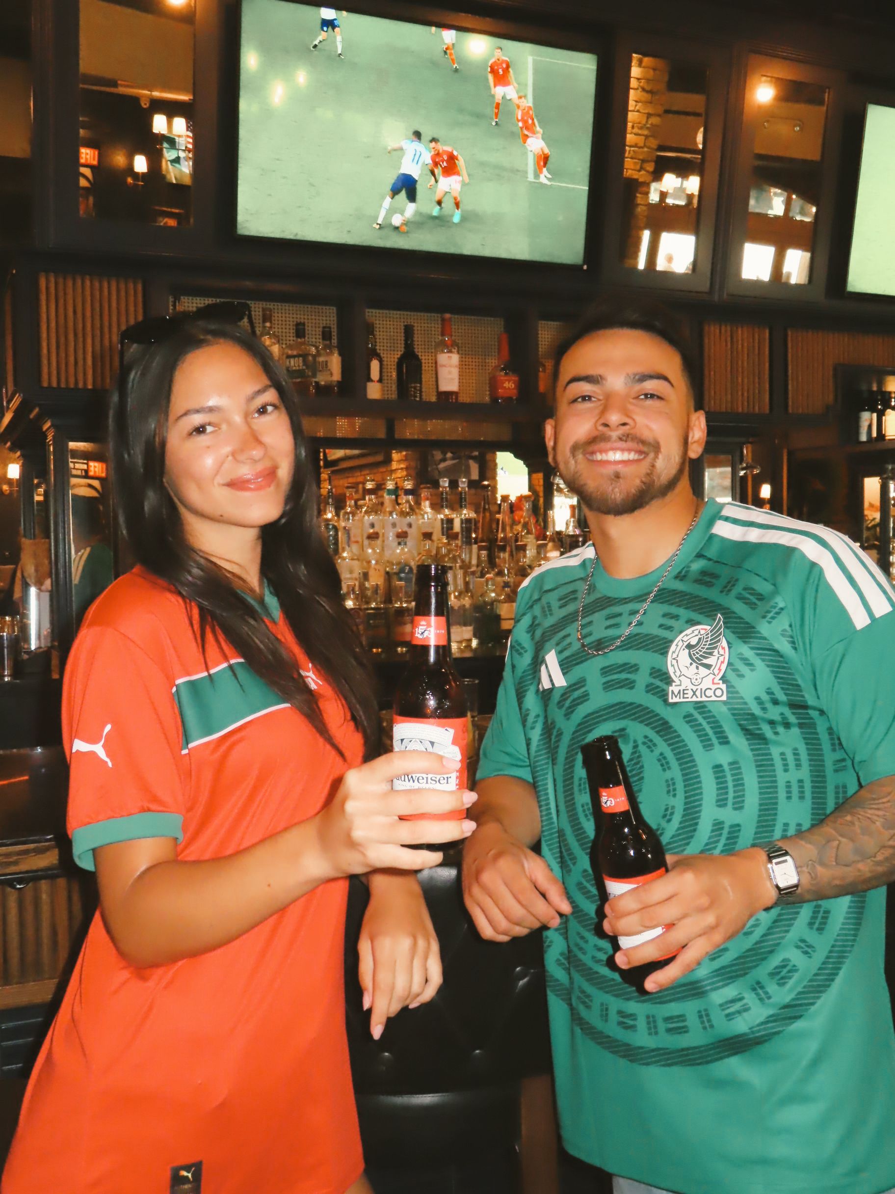 Two people in soccer jerseys smile and hold beer bottles inside a bar, catching the World Cup 2026 action on TV screens in the background.