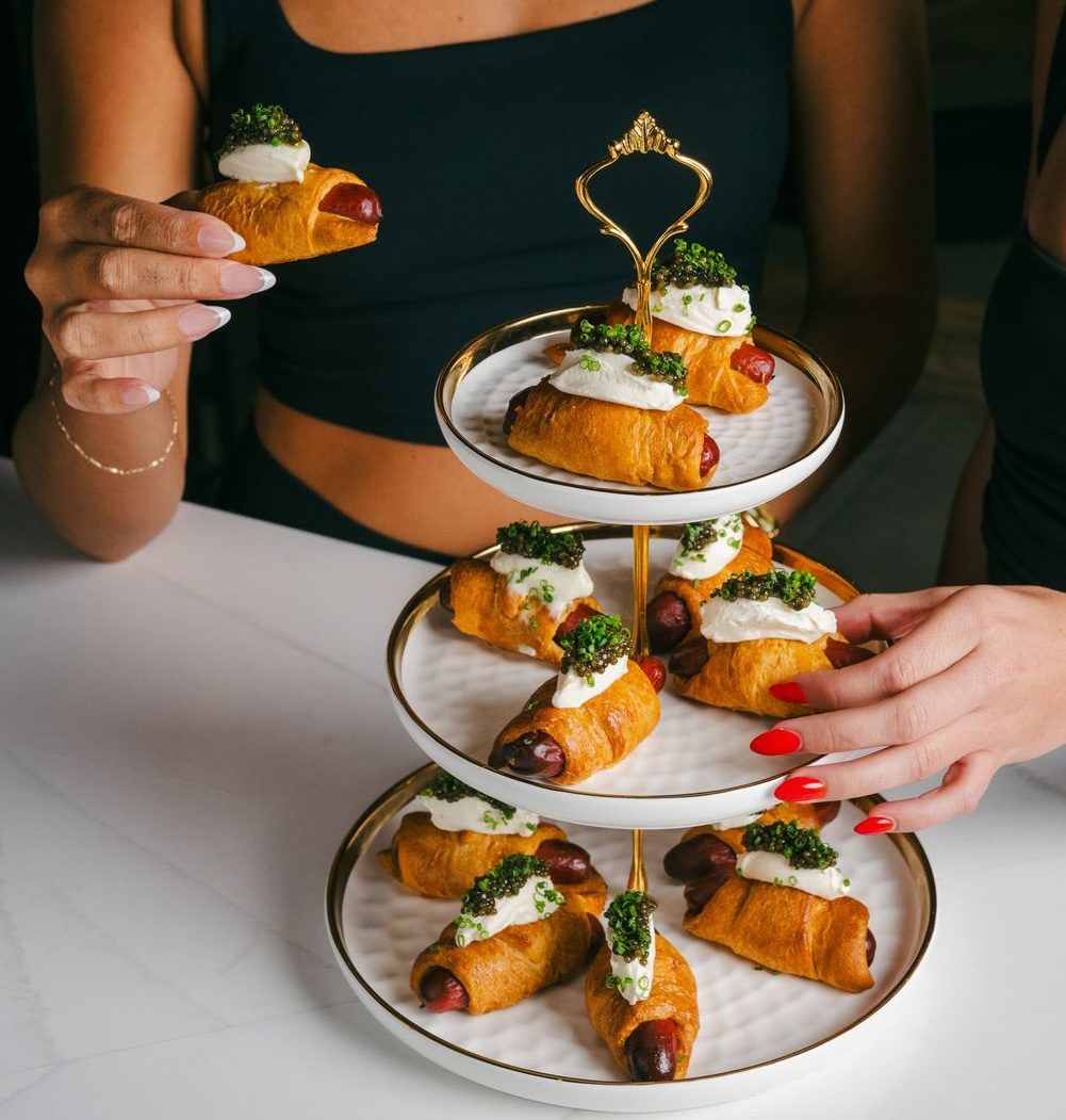 Two people hold pastry-wrapped sausages topped with cream and garnish, taken from a three-tiered serving tray filled with similar appetizers on a white table, perfect for enjoying during World Cup 2026 celebrations.