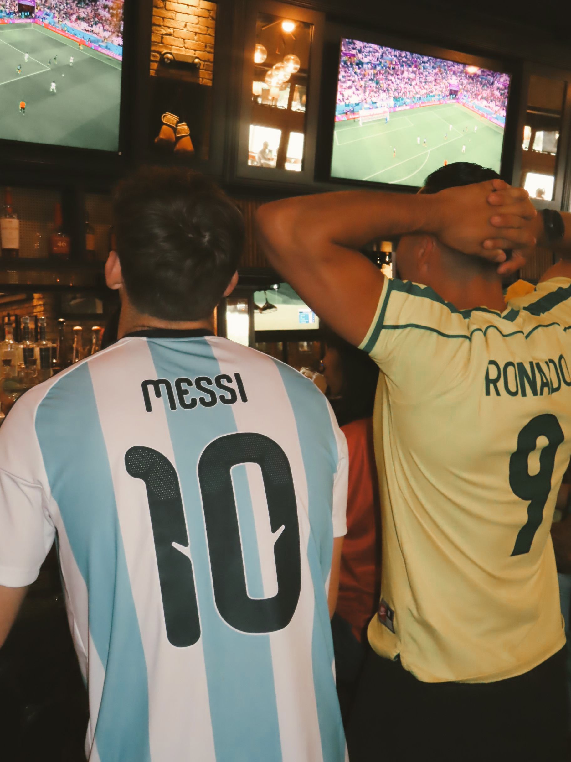 Two people at a bar watch soccer on TV screens. One wears an Argentina 