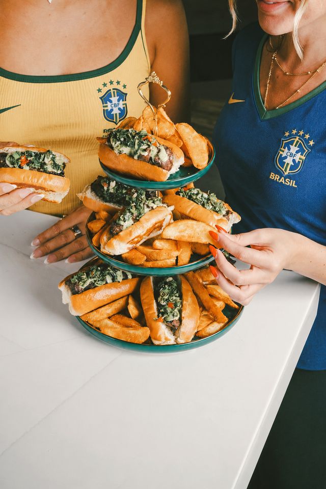 Two women in yellow and blue Brazil soccer jerseys hold a tiered tray with hot dogs topped with green relish and thick potato wedges, ready to eat at a white table while celebrating the excitement of World Cup 2026.