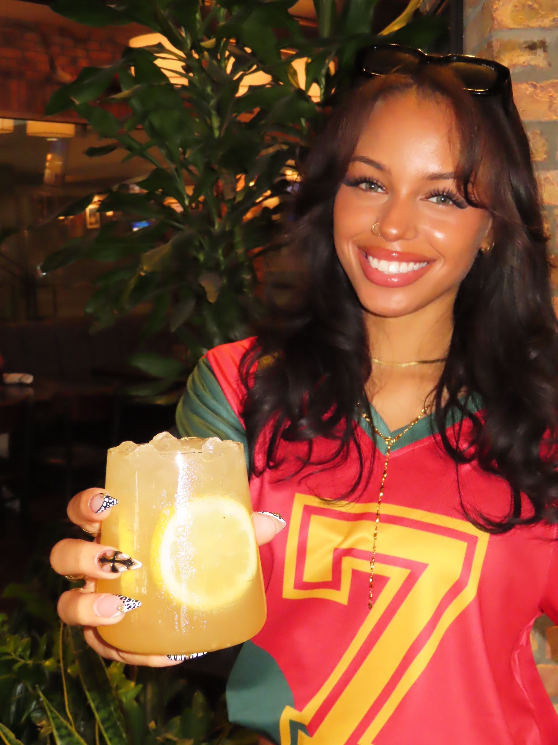 A smiling woman with long dark hair, wearing a red and green shirt with a yellow number 7, holds a glass of iced drink with a lemon slice. Gasparilla vibes shine through, with plants and a brick wall in the background.