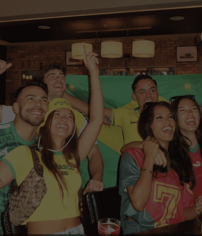 A group of six young adults cheer and smile in front of a Brazilian flag inside a bar, wearing colorful Brazil-themed clothing and football jerseys, celebrating together as they gear up for World Cup 2026.