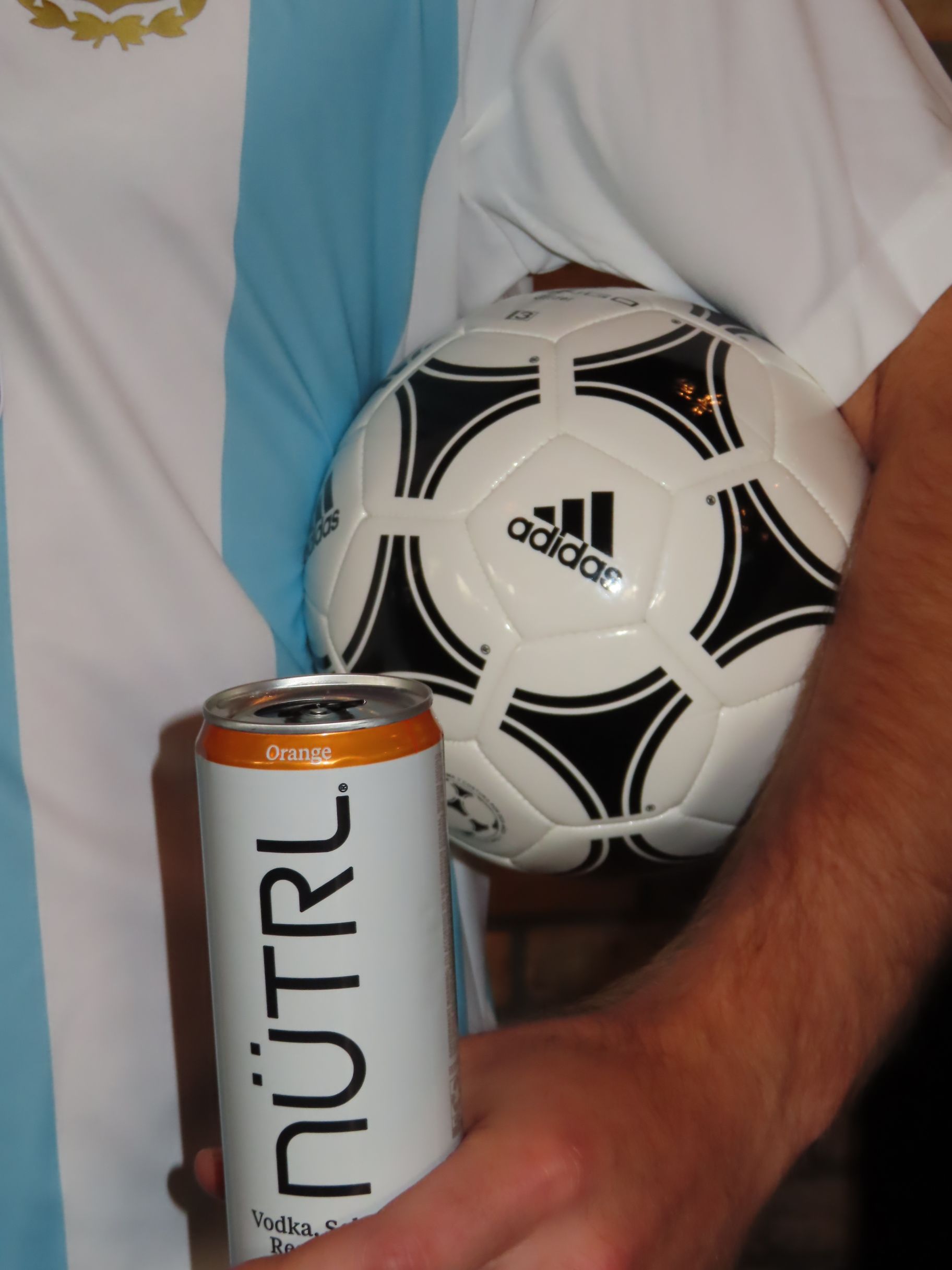A person in a light blue and white striped jersey holds an Adidas soccer ball under one arm and a can of NÜTRL orange vodka soda in the other, ready to celebrate Gasparilla in style.