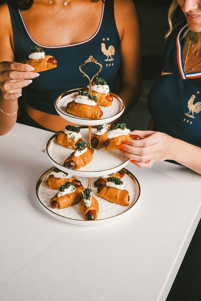 Two women in matching blue sports tops enjoy pastry-wrapped sausages topped with cream and herbs from a three-tiered tray, celebrating the spirit of World Cup 2026 at a white table.