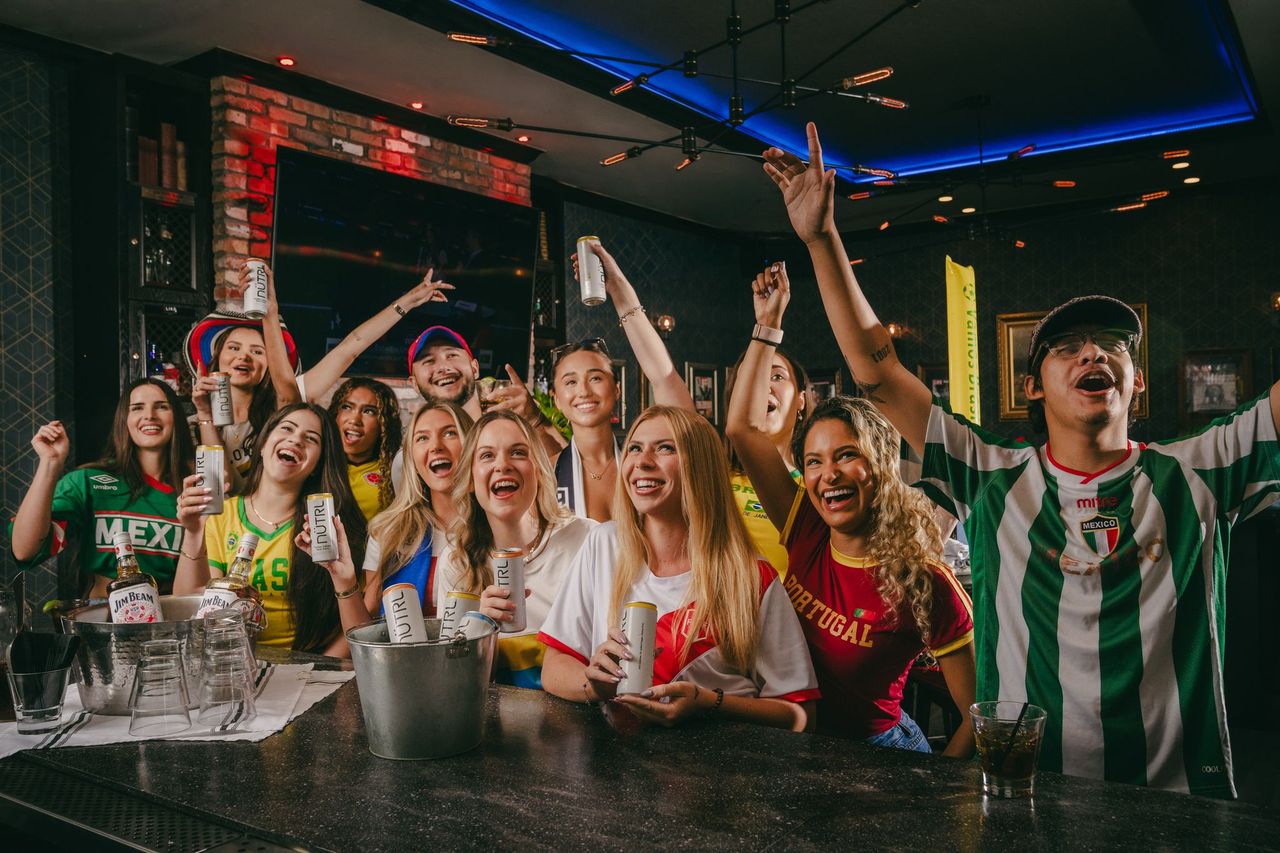 A group of enthusiastic people in soccer jerseys from different countries cheer and celebrate together at a bar, holding drinks and bottles, creating a lively and festive World Cup 2026 atmosphere.