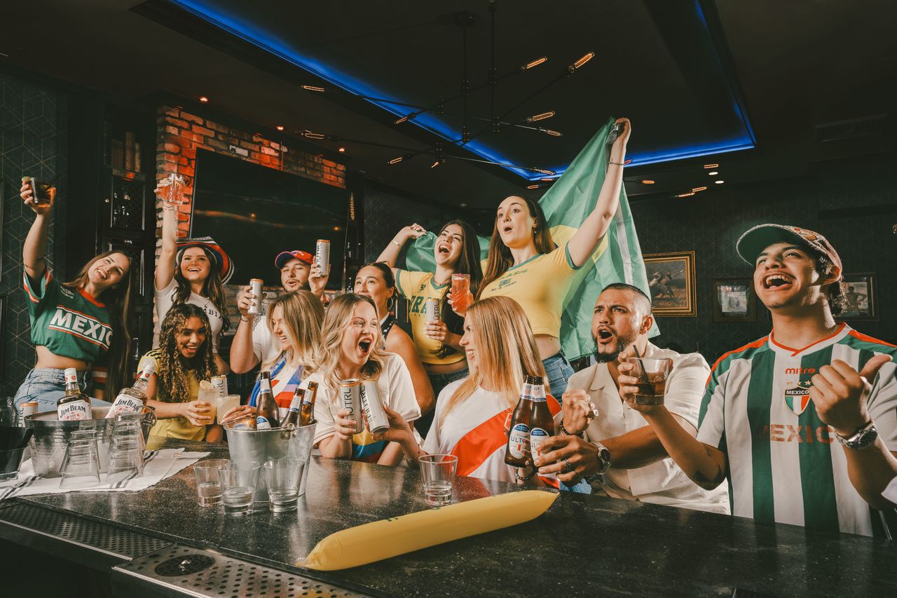 A group of excited people at a bar, wearing national soccer jerseys and holding drinks, cheer and celebrate together. Flags and team colors suggest they are watching a World Cup 2026 match.