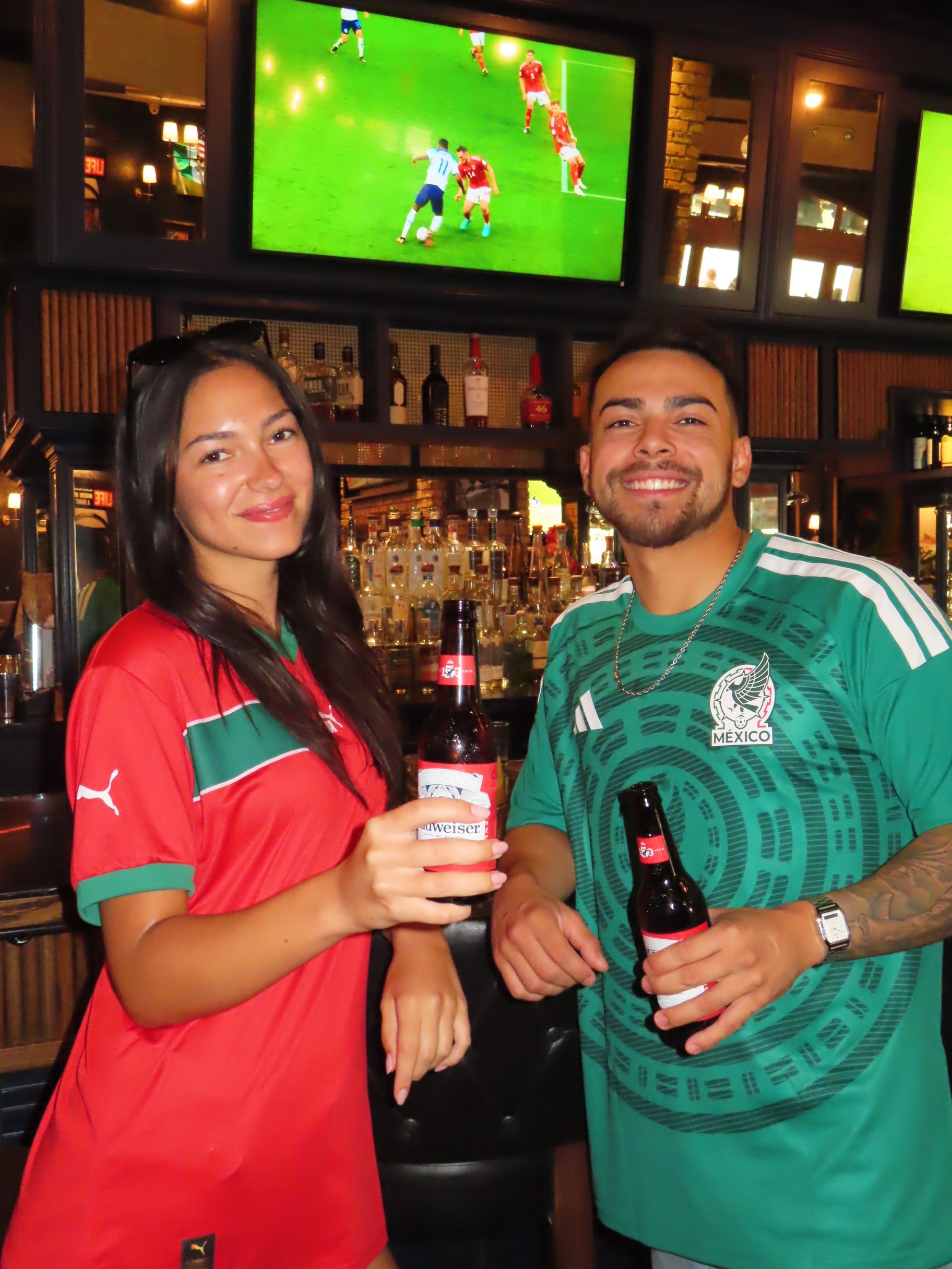 A woman and man smiling and holding beer bottles in a bar, both wearing soccer jerseys and festive Gasparilla beads. Televisions in the background display a soccer match.