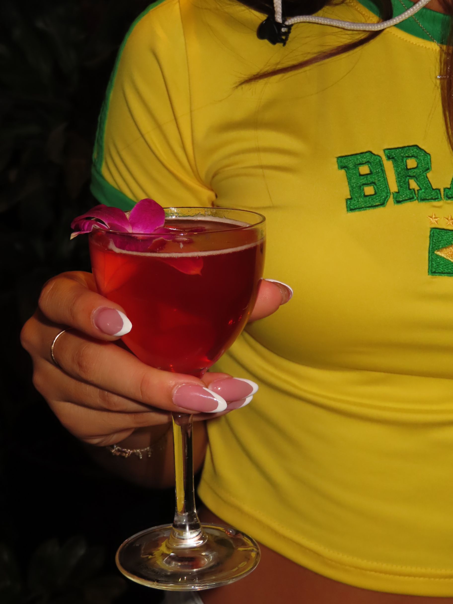 A person wearing a yellow Brazil jersey holds a festive gasparilla-inspired red cocktail garnished with a purple flower. Only their hand and upper body are visible.
