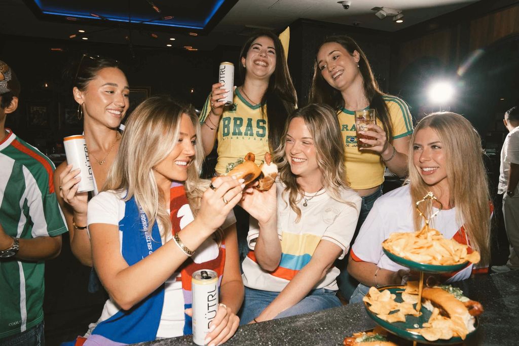 A group of six women wearing soccer jerseys and holding drinks smile and laugh together at a bar. Enjoying snacks like chips and pretzels, they appear to be celebrating or watching a World Cup 2026 match.