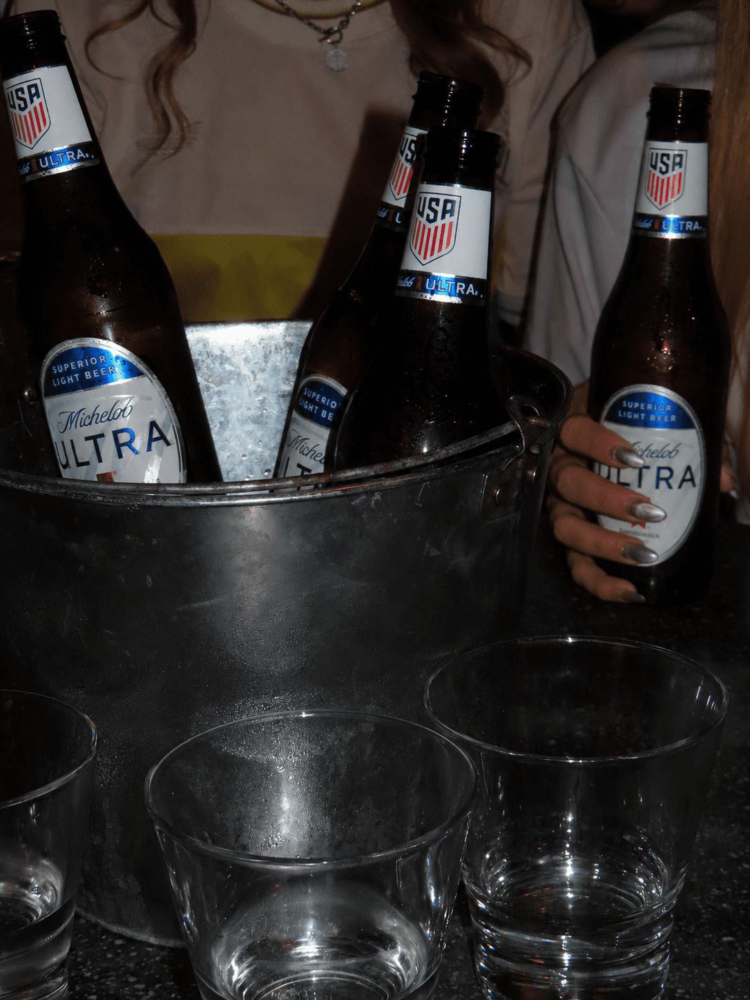 Four Michelob Ultra beer bottles, three in a metal ice bucket and one held by a person with painted nails, sit on a dark table with three empty glass tumblers—perfect for enjoying during the F1 Miami Grand Prix. Two people are partially visible in the background.